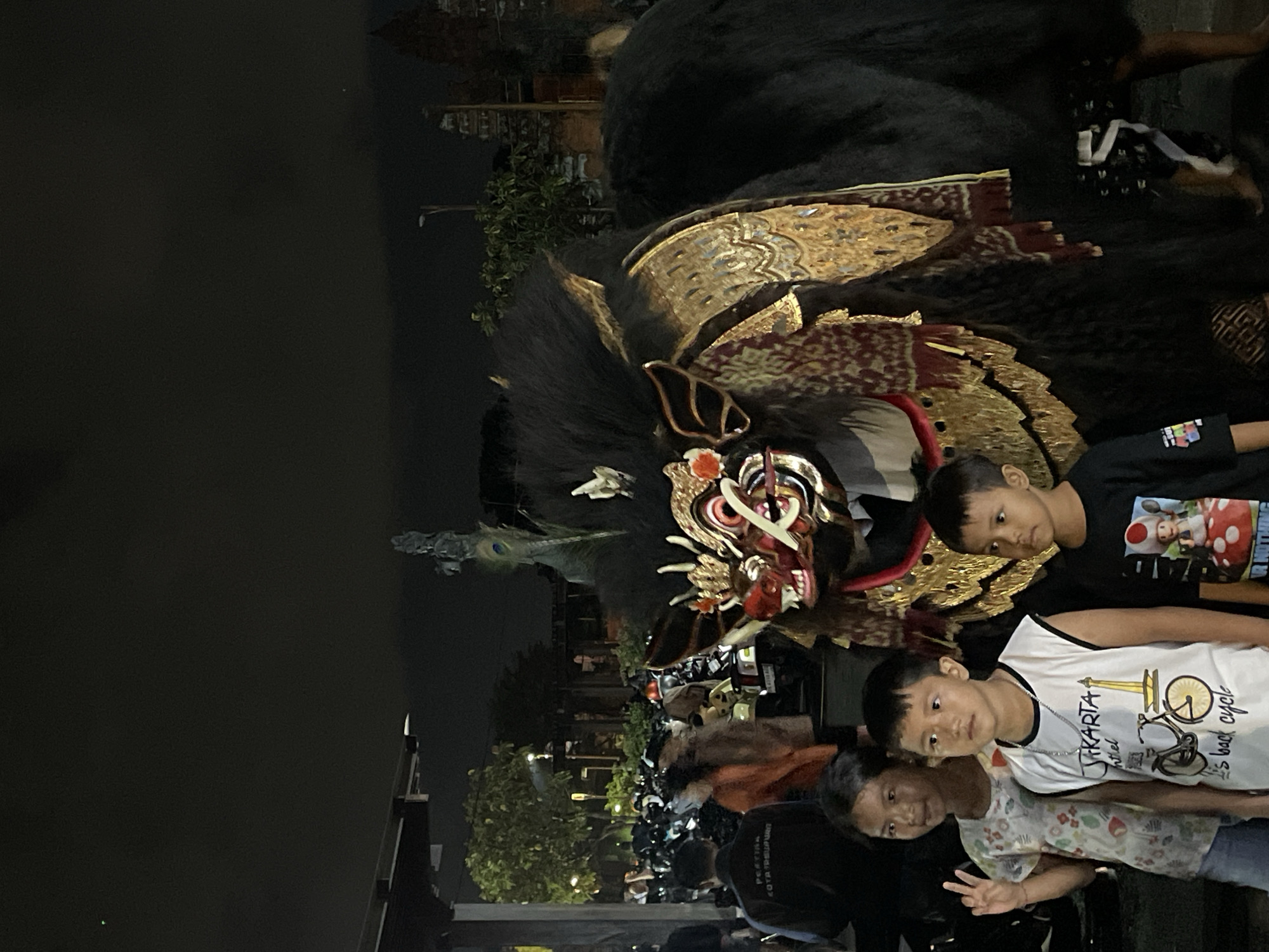 The image shows a group of people, including three young boys and at least one young girl, gathered outdoors at night. They are in front of a large, elaborate costume that appears to be a Barong, a mythical creature from Balinese mythology, often depicted as a lion-like beast with ornate golden headdress and intricate detailing. The Barong costume is adorned with gold and red patterns, and a peacock feather tail is visible.
The scene suggests a cultural event or festival taking place in Denpasar, Indonesia. The presence of numerous motorcycles parked in the background indicates a public gathering. The lighting suggests it is nighttime, possibly illuminated by streetlights and the costume itself. The expressions on the faces of the children are curious and engaged, observing the Barong. The overall atmosphere seems festive and communal.
One of the boys is wearing a black t-shirt with an image of Super Mario and a mushroom. Another boy is wearing a white sleeveless shirt with "JAKARTA" and "it's back cycle" printed on it, along with a bicycle graphic. There is also visible text on the back of a dark shirt worn by someone in the foreground, reading "PERTA" and "KOTA SEMPURNA".
