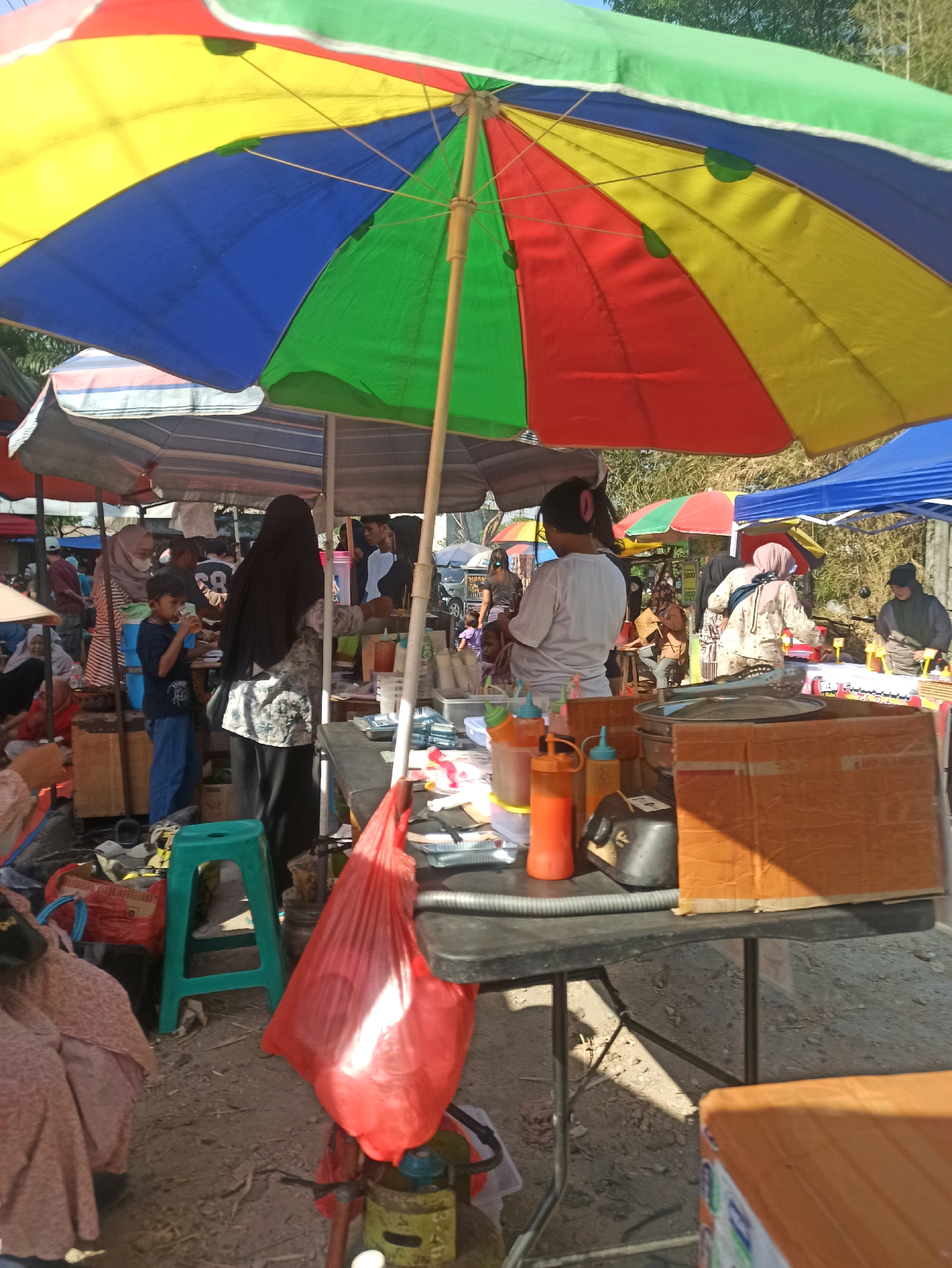 The image depicts a busy outdoor market scene in Telagamurni, Indonesia, under a large, multi-colored umbrella providing shade. Several stalls are visible, with people engaged in various activities. In the foreground, a table is laden with food-related items, including squeeze bottles of sauces, containers, and a large cardboard box. A green plastic stool sits to the left of the table, and a red plastic bag hangs from it. A person in a black hijab is seen at a stall to the right, interacting with a customer. Another stall behind them has a blue awning and appears to be selling food. In the background, more stalls and people are visible, suggesting a bustling market environment. The ground is dirt and gravel, and trees can be seen in the distance, indicating an outdoor setting.