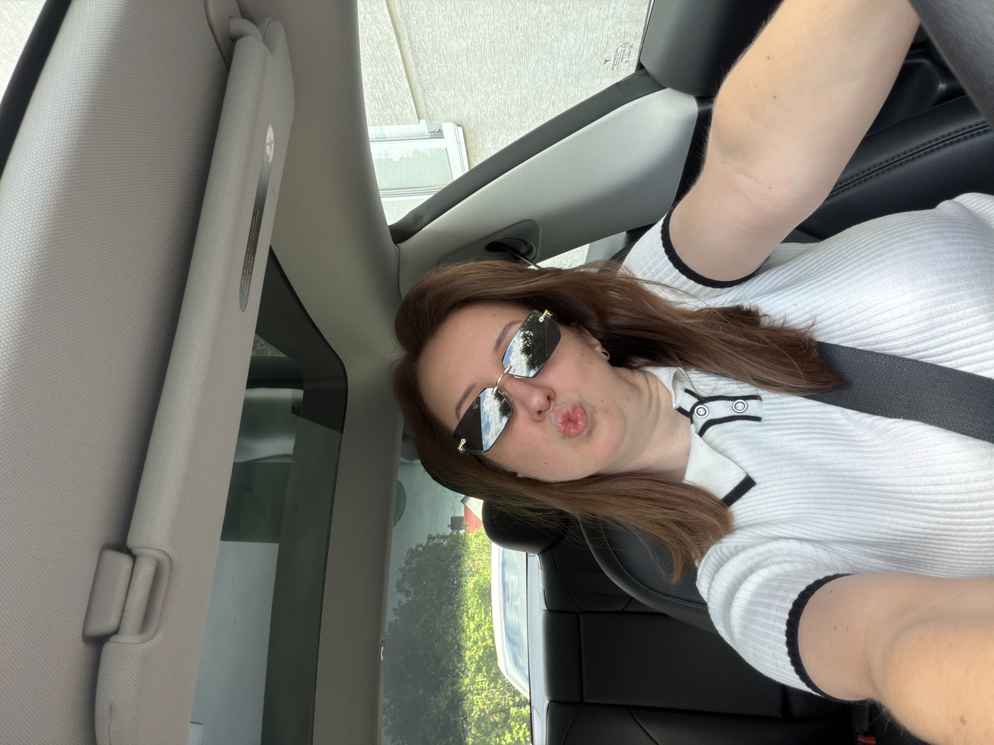 A woman is sitting in the driver's seat of a car, holding her phone up to take a selfie. She is wearing a white ribbed polo shirt with black trim and black-rimmed sunglasses. Her lips are pursed, as if she is blowing a kiss. The car has a panoramic sunroof, and the interior is black leather. Outside the car, there are trees and a white building visible. The photo was taken on a sunny day.