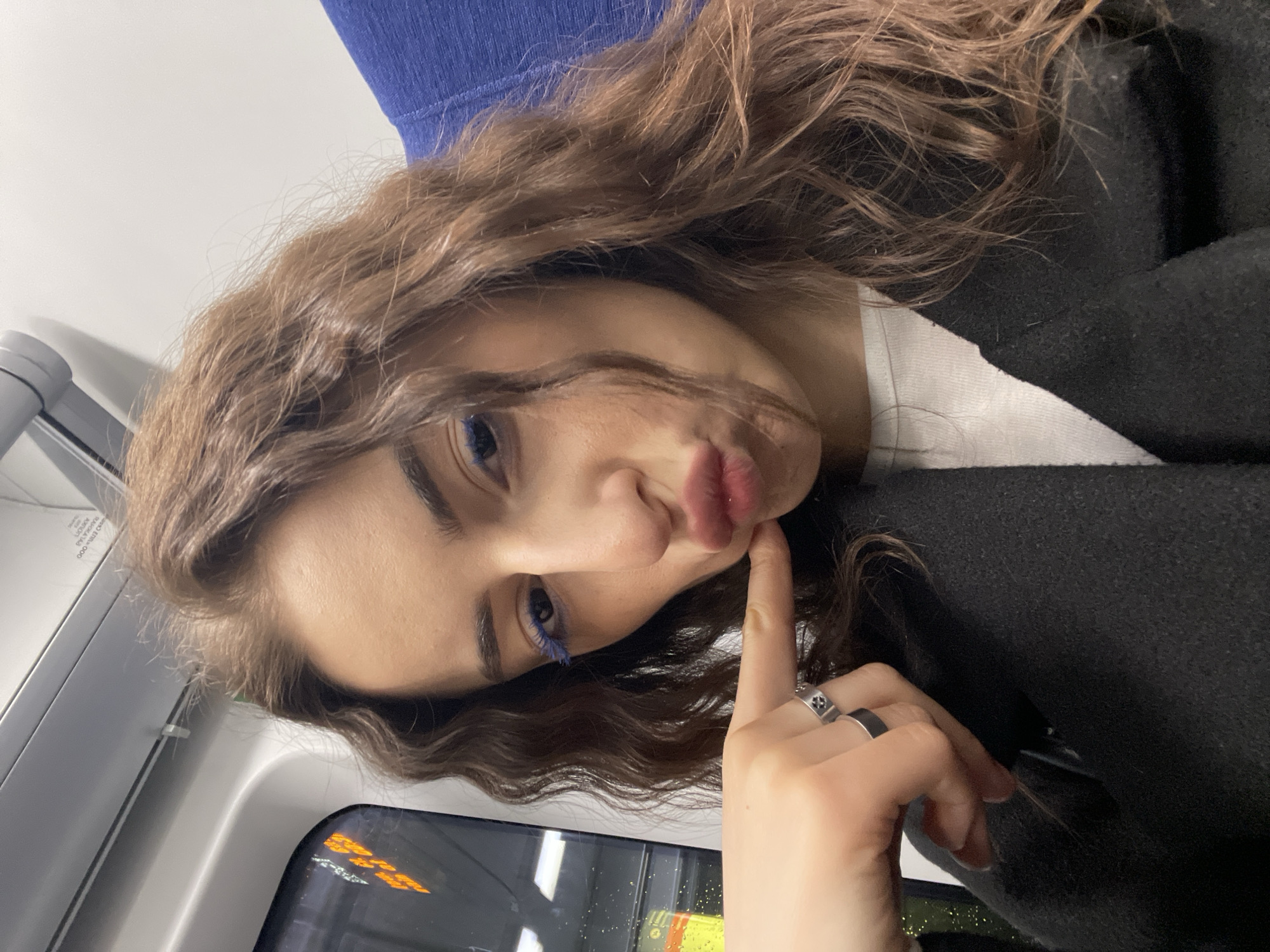 A young woman with brown curly hair, wearing blue mascara, is posing for a photograph. She is wearing a black jacket and a white top. Her right index finger is touching her lips. She is wearing two rings on her right hand. The photo appears to have been taken inside a train, possibly a Moscow Metro train, indicated by the blue seating and window. Orange lighting can be seen through the window.