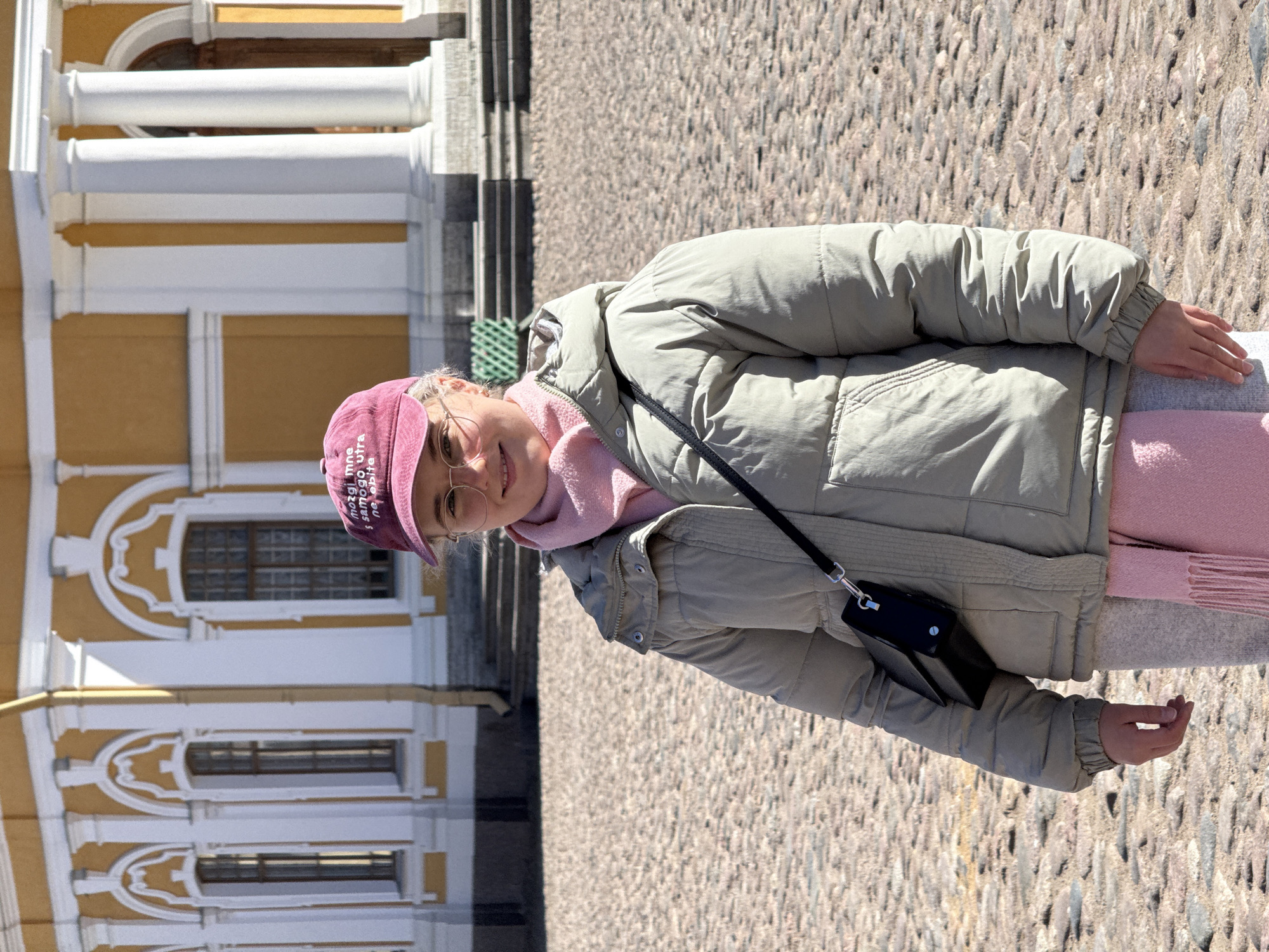 A full body portrait of a woman standing on a cobblestone street, with a large building in the background. The woman wears a beige puffer jacket, a pink scarf, and a dark pink baseball cap, along with wire-rimmed glasses. The building has a white facade with yellow accents, and features rows of windows. The location is the courtyard of Catherine Palace in Tsarskoye Selo, a part of the city of Saint Petersburg, Russia.
