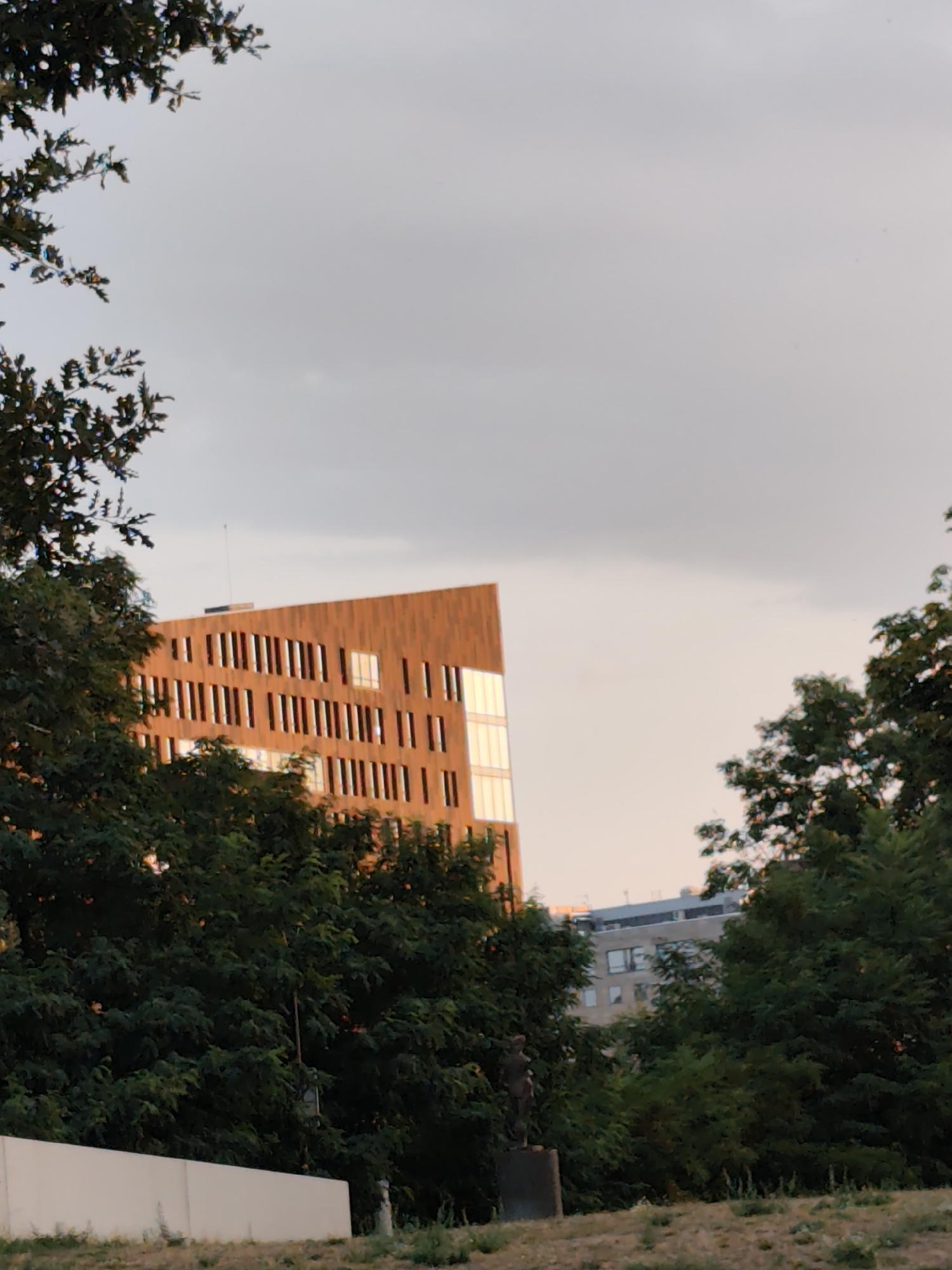 A low-angle outdoor shot captures a segment of a building with a facade of vertical wooden slats and regularly spaced windows, appearing to be under construction or recently built. The sky overhead is overcast. In the foreground, lush green trees partially obscure the view of the building and other structures. A hint of a statue is visible among the trees on the right side of the image. The scene suggests an urban park or a building site adjacent to green space.
