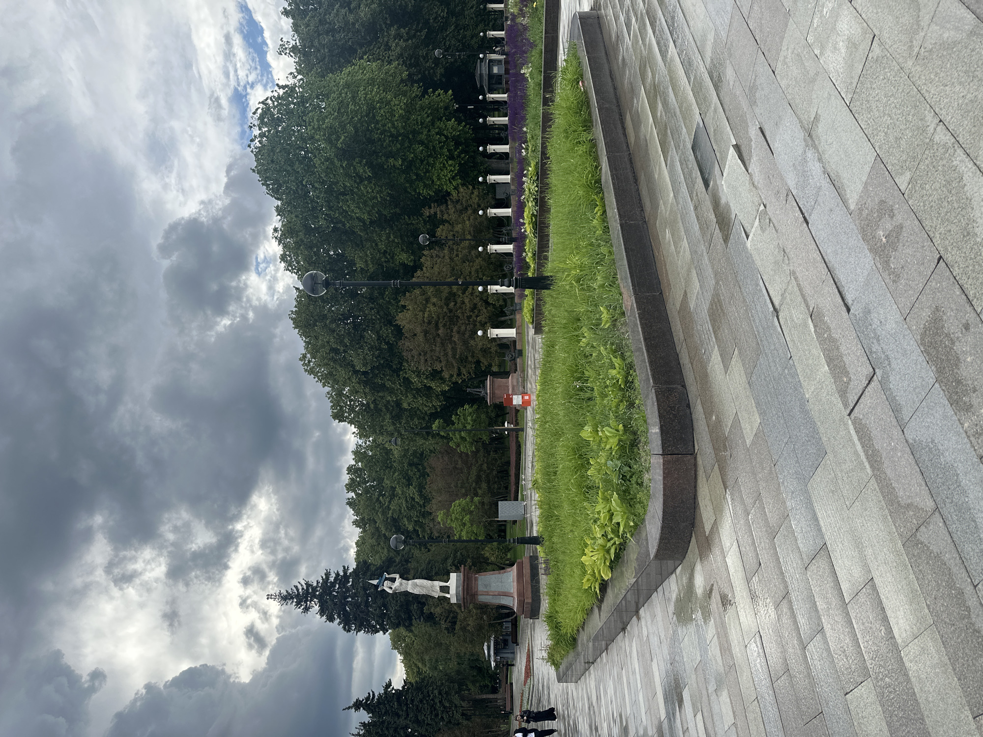 This media file captures an outdoor scene in what appears to be a park or public square, presented in a vertical orientation. The foreground is dominated by a stone pathway made of large, rectangular grey tiles, which are wet and reflective, suggesting recent rain. To the left of the pathway is a neatly manicured strip of vibrant green grass and lush foliage, bordered by a dark stone edge.
Beyond the green strip, a row of evenly spaced lampposts with spherical tops lines a path. In the background, a dense line of mature trees with dark green foliage forms a natural boundary. A few people can be seen in the distance, walking along the path.
Further into the scene, there is a prominent statue of a woman holding what looks like a flag or banner. The statue stands on a pedestal. Behind the statue and trees, the sky is filled with dramatic, heavy grey clouds, with some patches of brighter sky suggesting diffused sunlight. The overall mood is somewhat somber due to the cloudy weather, but the lush greenery provides a sense of life and nature. The time of day is unclear, but the overcast conditions suggest it could be daytime. There is no visible text in the image.