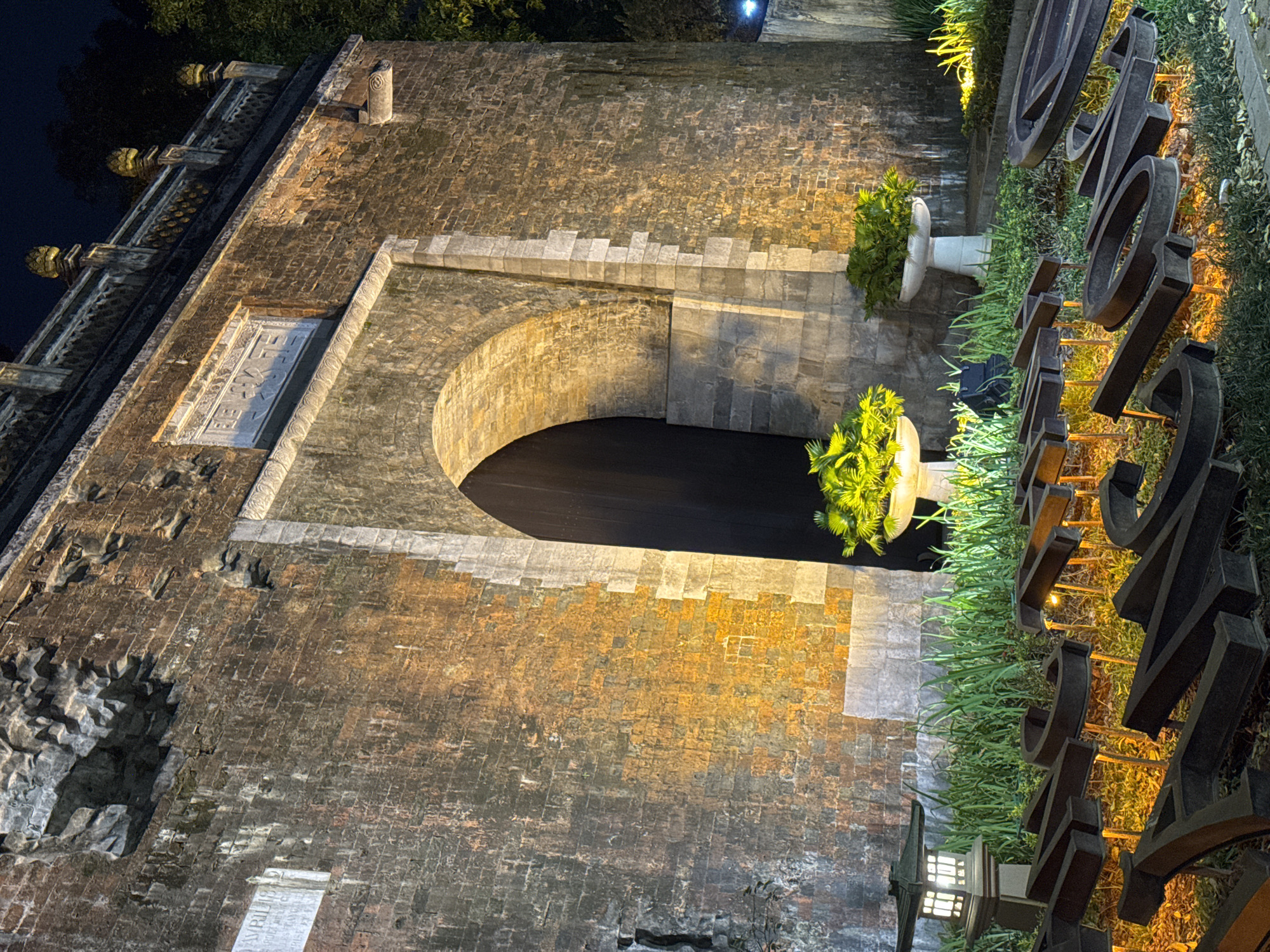 The image shows the entrance to the Imperial Citadel of Thang Long in Hanoi, Vietnam at night. The ancient brick gate is illuminated by warm spotlights, casting an orange glow on the weathered facade. Large stone blocks form the archway, which leads into a dark, cavernous interior. In front of the gate, ornamental plants in white planters sit amidst lush green grass. A traditional Japanese-style lantern stands to the left, adding a subtle, soft light to the foreground. Scattered on the ground are large, dark, three-dimensional letters, partially obscured by the grass and illuminated by small, yellow lights, spelling out "THANG LONG". The overall scene conveys a sense of history and grandeur, enhanced by the dramatic nighttime lighting.