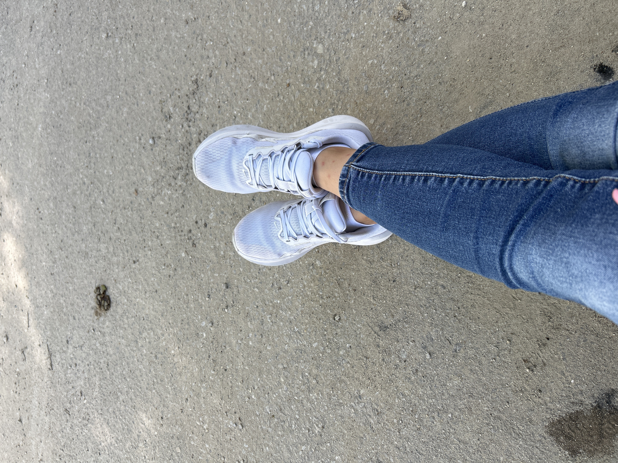 In a top-down perspective, a person's legs are crossed, clad in blue denim jeans. The individual is wearing white athletic shoes. The scene is set on a grey, textured surface, possibly asphalt or concrete, with small pebbles and debris scattered across it. In the upper left quadrant of the image, a small clump of organic material, possibly animal droppings, is visible on the ground. Sunlight casts shadows, suggesting an outdoor environment.