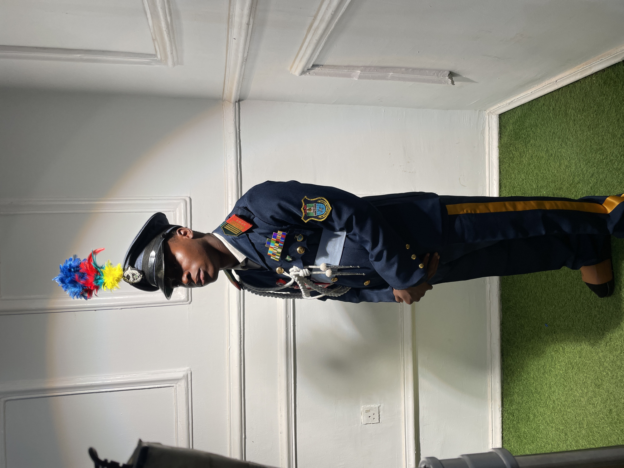 A person wearing a dark blue military uniform with gold trim, numerous medals, and a hat adorned with colorful plumes stands in a corner of a room. The walls are white with paneling, and the floor is covered in artificial green turf. The location appears to be indoors, possibly a studio or set. The person is positioned in the corner where the walls meet, with their body facing towards the right side of the frame. The setting does not reveal any specific location within the city of Barakin Akawo, Nigeria.