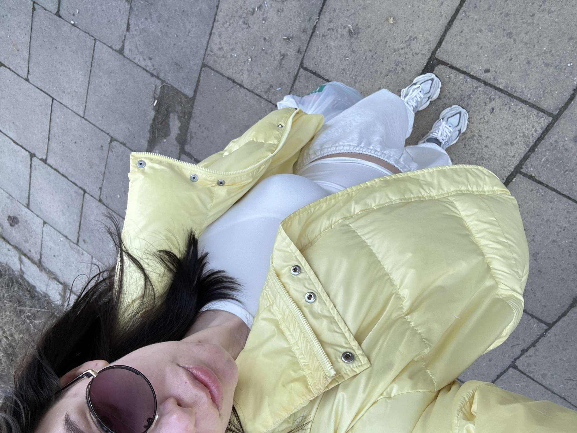 A young woman stands on a paved surface, possibly a sidewalk or plaza, likely in an urban setting based on the paving. She wears a light yellow puffer jacket, white pants, a white top, and white sneakers. She is also wearing round sunglasses with a dark lens. The pavement consists of large, rectangular gray stones. A plastic bag is partially visible near her feet. The woman is looking up towards the camera, taking a selfie, her dark hair partially obscuring her face.
There is no verifiable information about the location being the city of Chaikovsky, Russia.