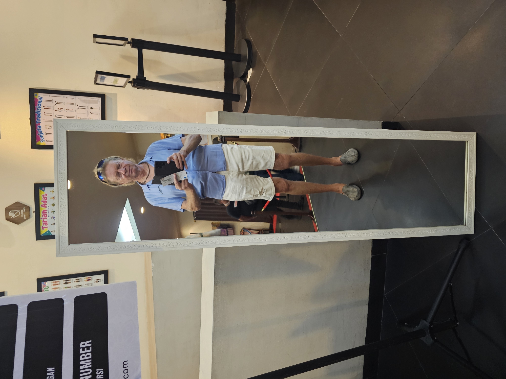 A man is taking a selfie in a mirror. The man is wearing a light blue button-down shirt, white shorts, and grey shoes. He is holding a smartphone in his right hand and a paper in his left hand. The mirror is tall and rectangular with a white frame. It is standing on a black tripod. The background includes signs and posters, and part of the decor of a shop in Kuta Selatan, Indonesia.