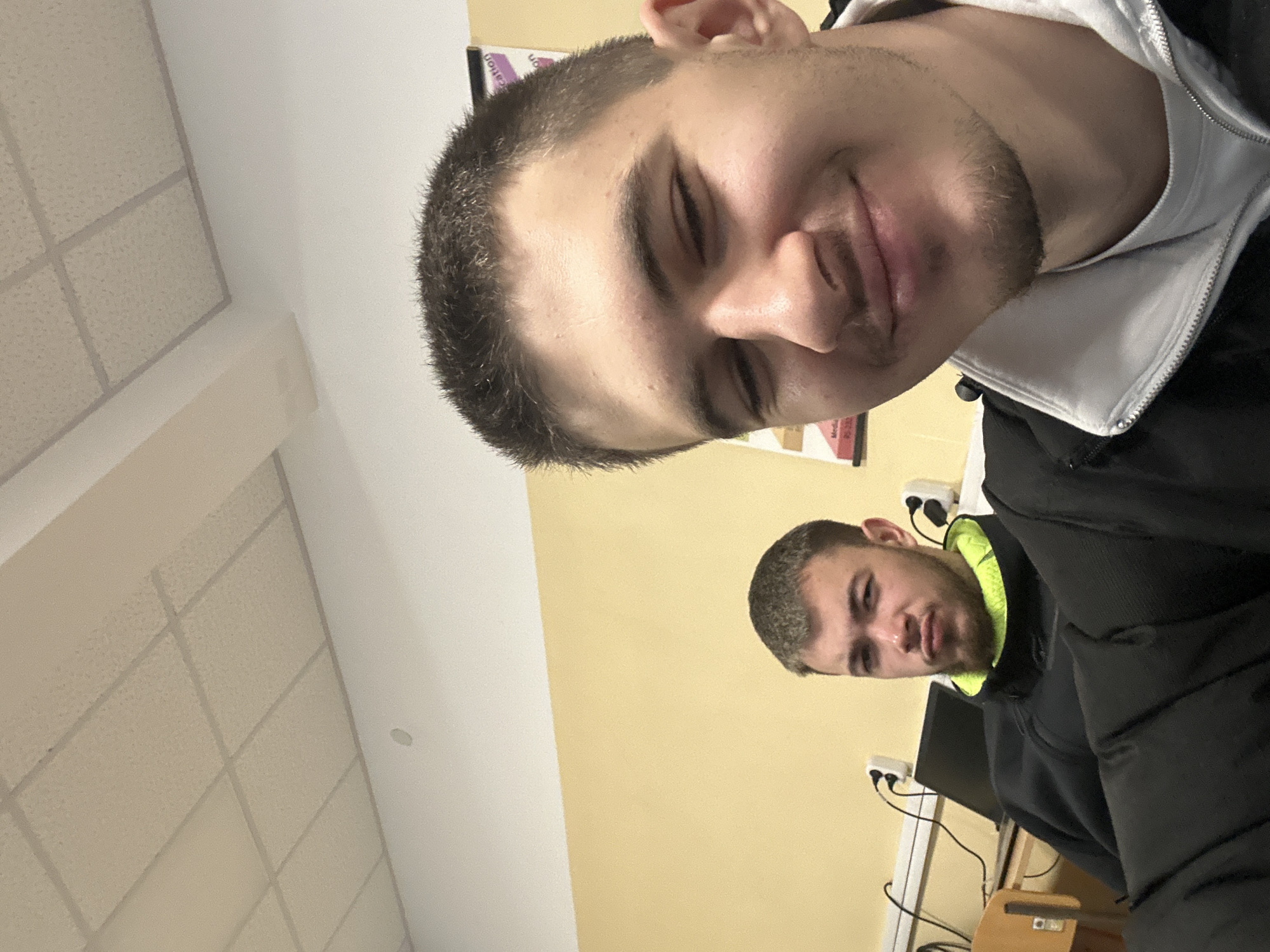 The image shows two men indoors. The first man is in the foreground, taking a selfie. He has short brown hair, a light complexion, and is wearing a white hoodie under a black jacket. The second man is in the background, slightly out of focus. He has short brown hair and a short beard, and is wearing a black jacket with a lime green collar. The background consists of a beige wall, electrical outlets, and what appears to be a computer monitor and desk. The ceiling is visible in the upper left corner, featuring square ceiling tiles.
The image does not contain enough information to determine the location in Sofia, Bulgaria.