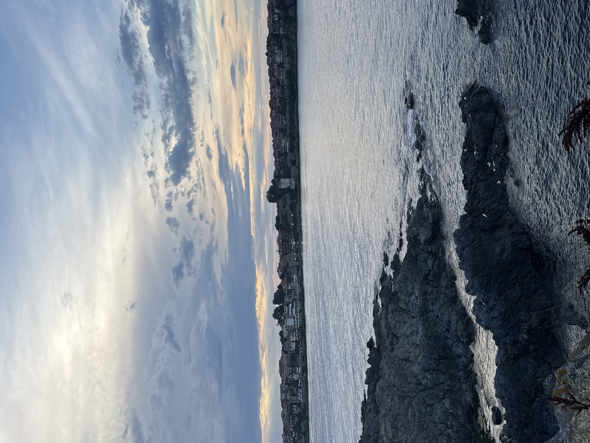 The image depicts a coastal view of the city of Sozopol, Bulgaria, during what appears to be dusk or early morning. In the foreground, dark, rugged rocks line the edge of the water, with some waves gently lapping against them. The sea itself is a deep blue-gray, reflecting the muted tones of the sky.
Across the water, the cityscape of Sozopol is visible, characterized by a dense collection of buildings with red-tiled roofs. The architecture suggests a historic European coastal town. The buildings are situated on a gentle incline, rising from the shoreline.
The sky above is filled with a mix of clouds. Towards the horizon, there are streaks of pale yellow and orange, indicating the presence of the sun, either just risen or about to set. Higher up, the clouds are a mix of gray and white, with some areas appearing brighter where the light is breaking through. The overall atmosphere is calm and serene. There are no people visible in the image. The environment is natural and urban, with the sea and rocks contrasting with the man-made structures of the city.