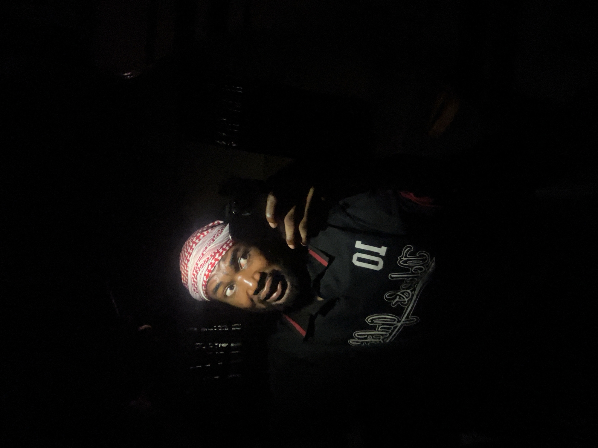The image shows a person wearing a red and white checkered headscarf and a black shirt with the number "10" on it. The person appears to be in a dimly lit environment, possibly indoors, as the background is dark and indistinct. No specific location in Lokoja, Nigeria, is identifiable. There are no discernible objects or interactions to describe beyond the person's presence.