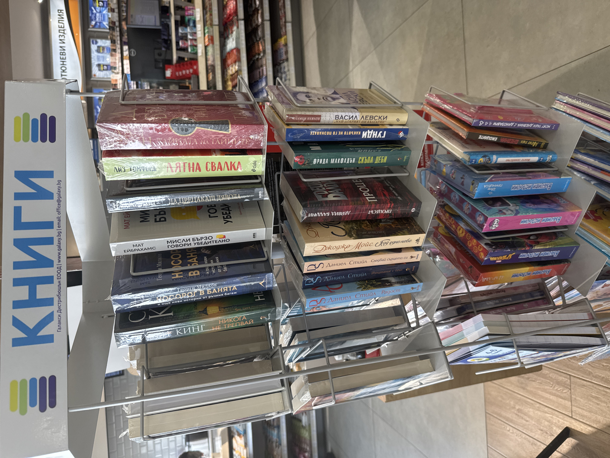 A close-up, slightly tilted, indoor shot shows three racks of books in a bookstore or retail environment. The racks are made of light gray wire. A display stand on the left has the word "КНИГИ" in large blue letters stacked vertically and the logo of "Галакси Дистрибюшън ЕООД" with a website and email address. The books on the left rack include titles such as "ЛЯТНА СВАЛКА" and "НОСОРОГ В БАНЯТА". The books on the middle rack contain titles such as "ВАСИЛ ЛЕВСКИ" and "ПРОЦЕСЪТ". The rack on the right has titles such as "БЪБРИВИ ИСТОРИИ". The setting appears to be a bookstore or retail area with other merchandise visible in the background. The location is implied to be within Bulgaria, based on the Bulgarian text. There is no information to suggest the city of Antimovo, Bulgaria. The floor is light-colored tile.