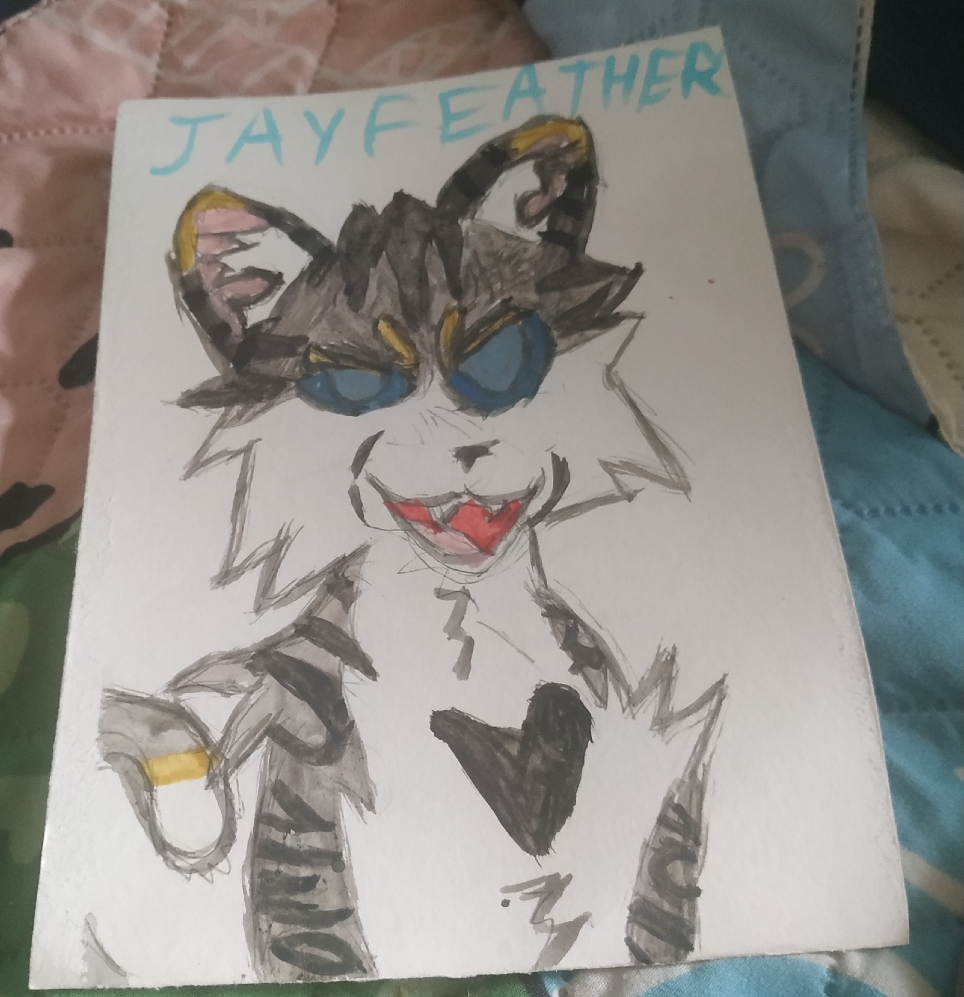 A drawing of a cat-like character labeled "JAYFEATHER" is on a piece of paper resting on a bed with multi-colored bedding. The character has gray and black fur, blue eyes, and an open mouth revealing red inside. It has striped markings and is holding a ring. The location is not provided and can not be derived from the image.
