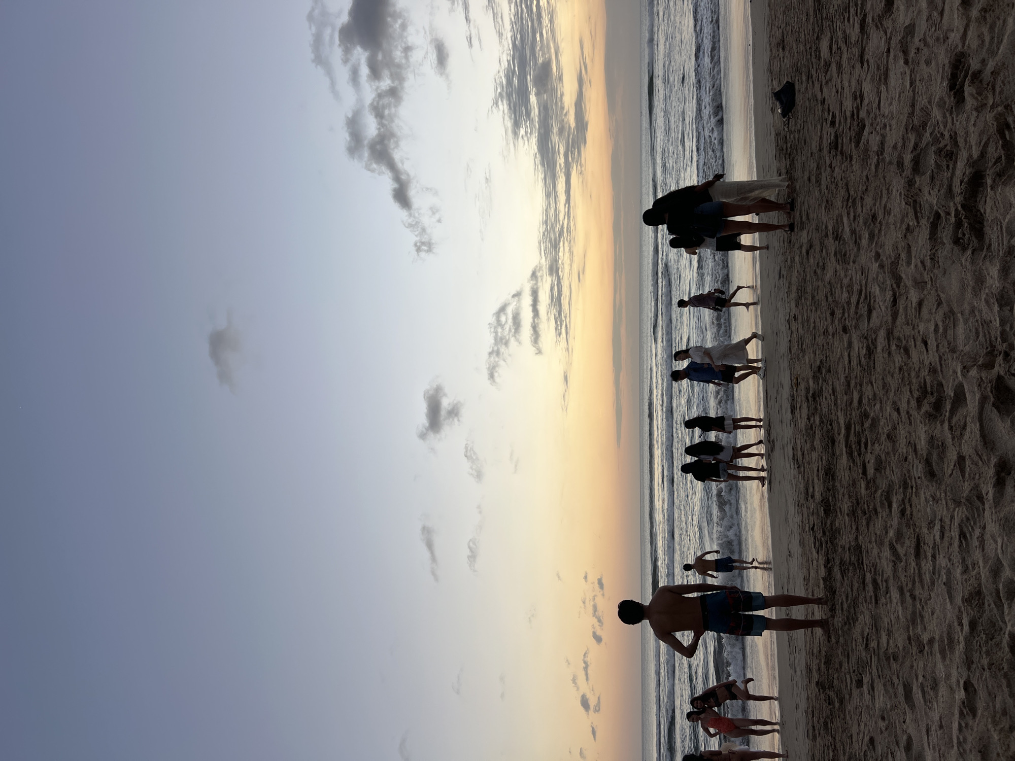 At sunset in Kuta, Bali, Indonesia, a group of people are gathered on a sandy beach. The sky exhibits hues of pale blue, soft orange, and yellow, with scattered clouds. The ocean, with gentle waves, stretches towards the horizon. Silhouettes of individuals are visible on the wet sand near the water's edge, some walking, others standing. In the foreground, the sand is textured and marked by footprints. The overall atmosphere is serene and indicative of evening leisure activities.