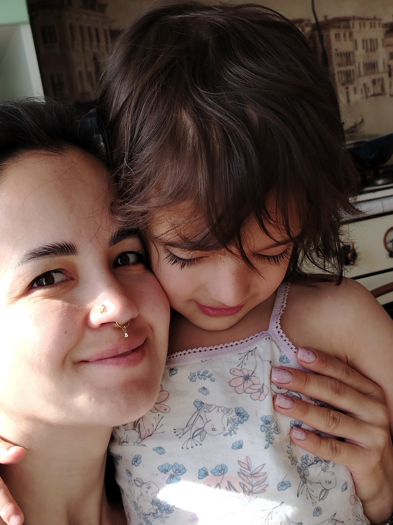 The image presents a warm, close-up portrait of two individuals, an adult woman and a young child, sharing an intimate moment indoors. The adult, positioned on the left, has dark hair, dark eyes, and distinct eyebrows. She wears two nose piercings: a gold septum ring featuring a small dangling diamond and a gold nostril stud with a cross or star design. A gentle smile graces her lips as she looks towards the camera, with bright natural light highlighting her face.
The child, nestled close to the adult on the right, has dark, wavy hair and long eyelashes. Her gaze is directed downwards, suggesting shyness or contemplation, as she leans into the adult. She is dressed in a light-colored top adorned with a subtle pattern of blue flowers and bunnies, finished with light purple lace trim. The child's left hand, with light pink, possibly glittery, nail polish, is gently placed on the adult's arm or shoulder, conveying affection.
The background, softly blurred, features wallpaper depicting an architectural scene reminiscent of old European cities, with detailed buildings and hints of canals or waterways. A portion of a white appliance or cabinet is visible on the far right. The strong natural light suggests it is daytime. The overall ambiance is one of tenderness and a close familial bond. There is no visible text in the image. The context provided indicates the location is Noviy, Russia.