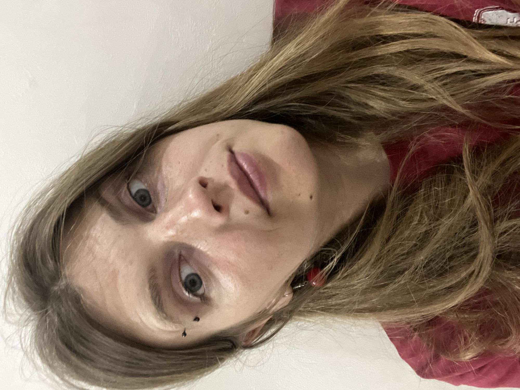 A close-up, head-and-shoulders selfie of a young person with long, blonde hair, taken indoors against a white wall. The person has blue eyes, a nose piercing, and is wearing a red shirt. They are also wearing a red, heart-shaped earring. The lighting is soft, and the focus is on the person's face. The location is not specified within the city of Сеніцкі сельскі Савет, Belarus, and no context is provided for the image.