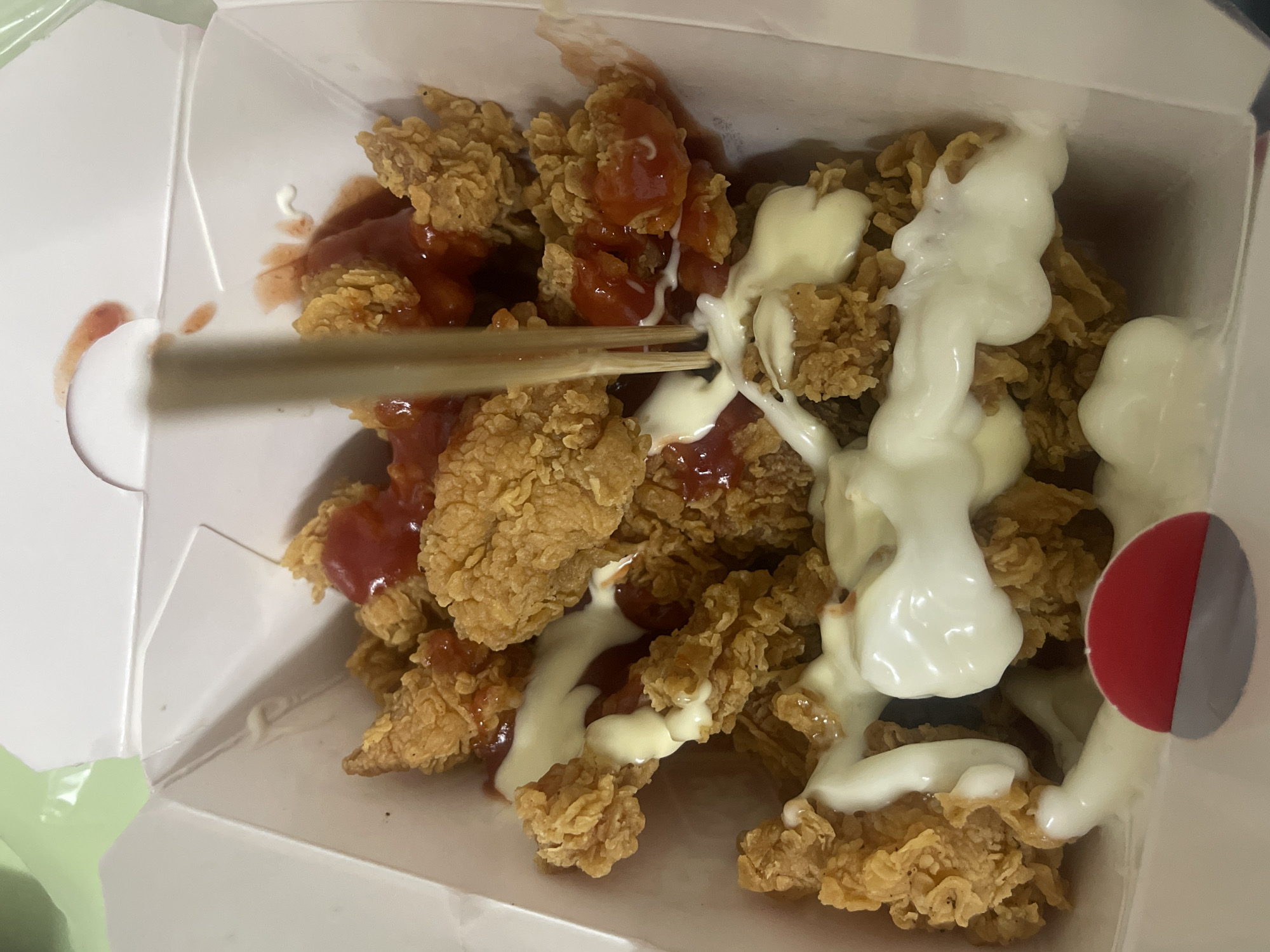 The image shows a white take-out box filled with fried chicken pieces covered in red and white sauces, being picked up by wooden chopsticks. The location is Chongqing High-tech Zone, China. No people are visible.