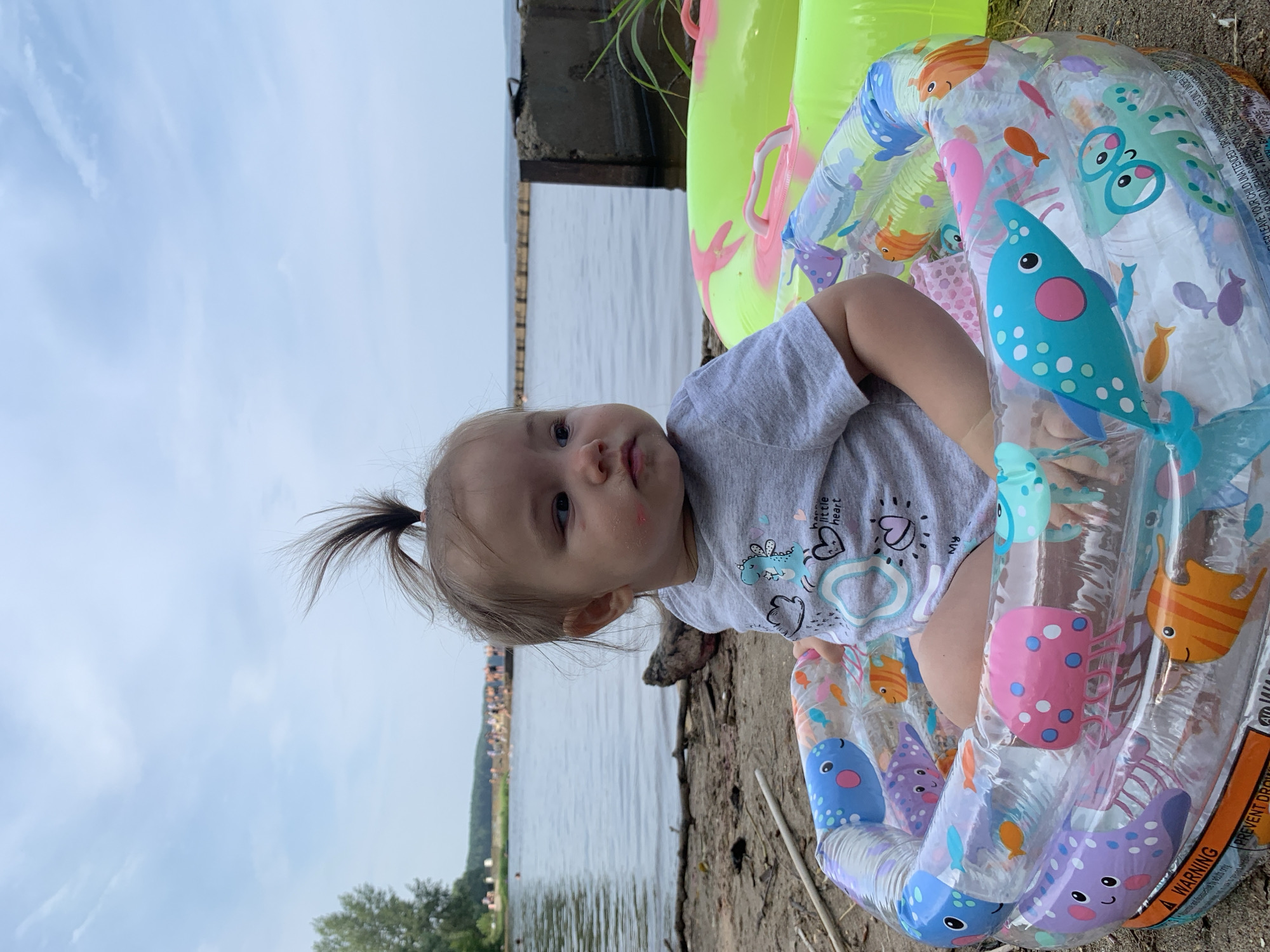 A young child is sitting in a small, inflatable kiddie pool decorated with cartoon sea creatures. The child, facing to the right, has light brown hair tied in a high ponytail and is wearing a gray t-shirt with a dinosaur design. Red smudges are visible on the child's left cheek. The kiddie pool is filled with water and some toys. Behind the child, a large, bright green inflatable ring with pink accents is visible. The scene is set on a sandy, pebble-strewn shore next to a body of water, likely a river or lake. In the background, a shoreline with trees and a line of people can be seen, suggesting a public beach or recreational area. The sky is overcast with scattered clouds. The location appears to be the Kama River embankment in the city of Chaikovsky, Russia.