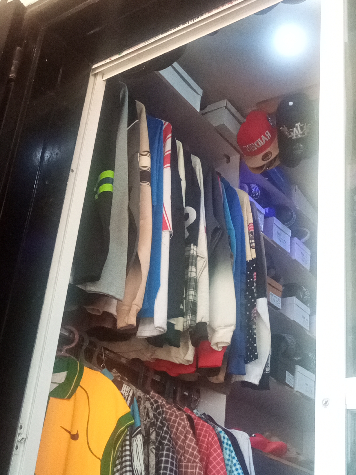 An indoor view of a retail space, potentially a clothing store, in Bukuru, Nigeria. The frame is filled with garments hanging on a clothes rack, with additional inventory stored on shelves in the background. Items include sweaters, shirts, and at least one yellow Nike polo. Some items appear to be new and packaged in boxes on the shelves above the hanging clothes. The space is illuminated by a light source, and the entrance or exit appears to be near the edge of the frame.