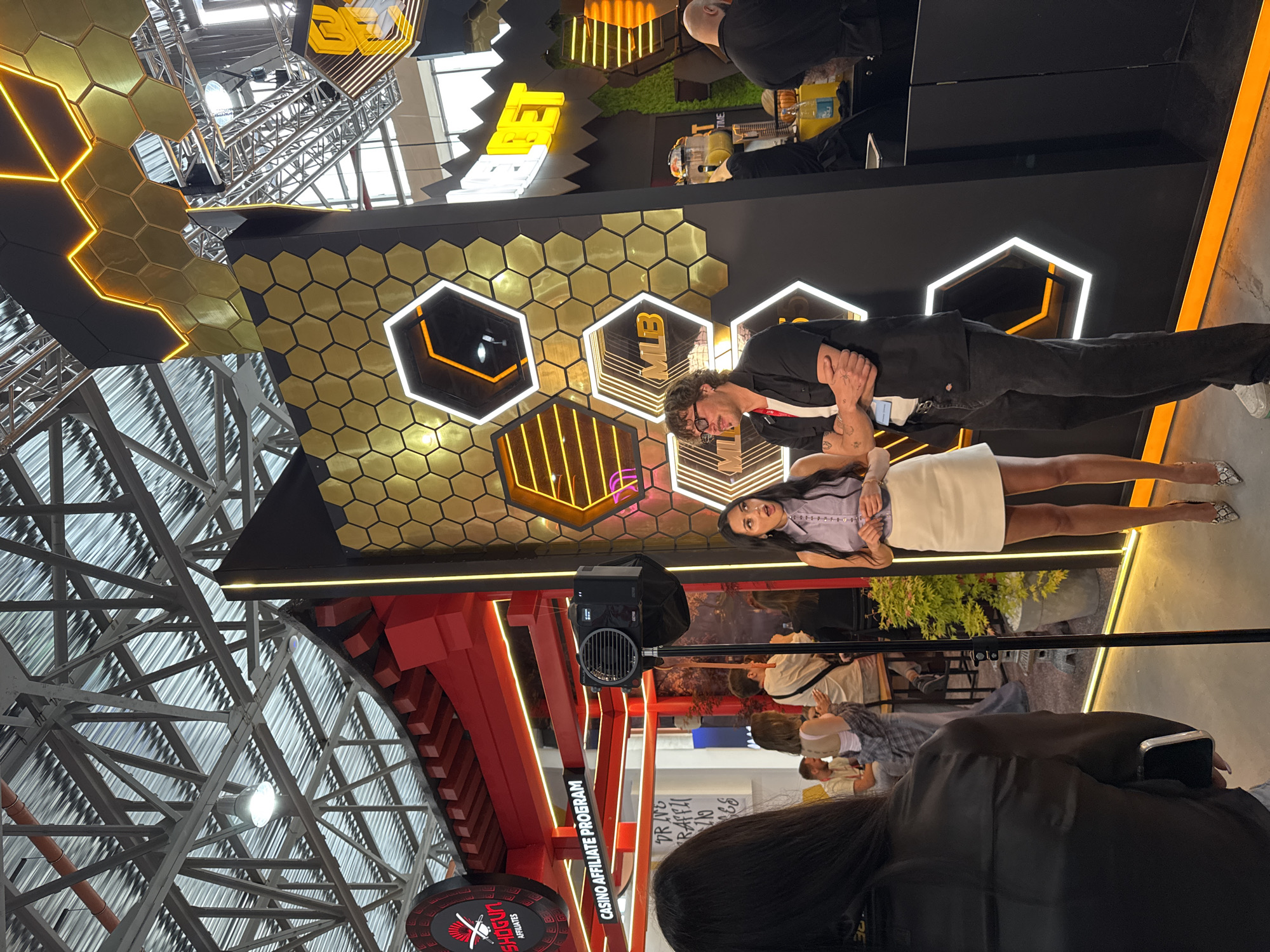 Captured indoors, the image presents a scene with two individuals standing in front of a backdrop consisting of interconnected gold and black hexagons, some illuminated with neon lights. One hexagon distinctly features the letters "MLB" in white neon.
The individuals comprise a woman and a man. The woman, on the left, wears a lilac blouse and a short white skirt paired with reptile-patterned heels. The man, to her right, is dressed in a dark suit. He has glasses on and his arms are crossed.
The environment includes elements of what seems to be a trade show or promotional event, indicated by a "Casino Affiliate Program" sign in red and white. A speaker with a black cover is situated in the center. A glimpse of greenery and other individuals can be seen behind the two primary subjects. Overhead, a complex metal structure supports the venue's roof.
The exact location of the image within Yerevan, Armenia cannot be identified from the visual data alone.