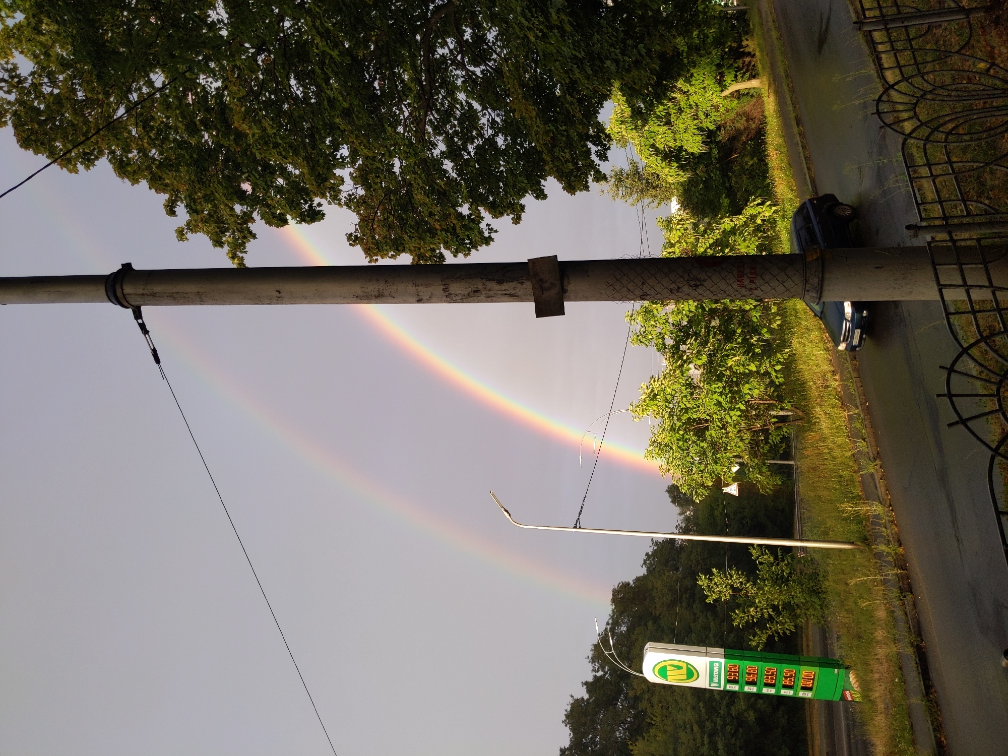In this image, a double rainbow arcs across a grey sky. A light pole stands prominently in the foreground, with a dark-colored car driving on a wet road. To the left, a green gas station sign displays fuel prices, including 93.80, 96.80, 83.50, and 85.90. Trees with lush green leaves surround the scene, and a black metal fence is visible at the bottom of the frame. The overall environment suggests a roadside scene after a rain shower.