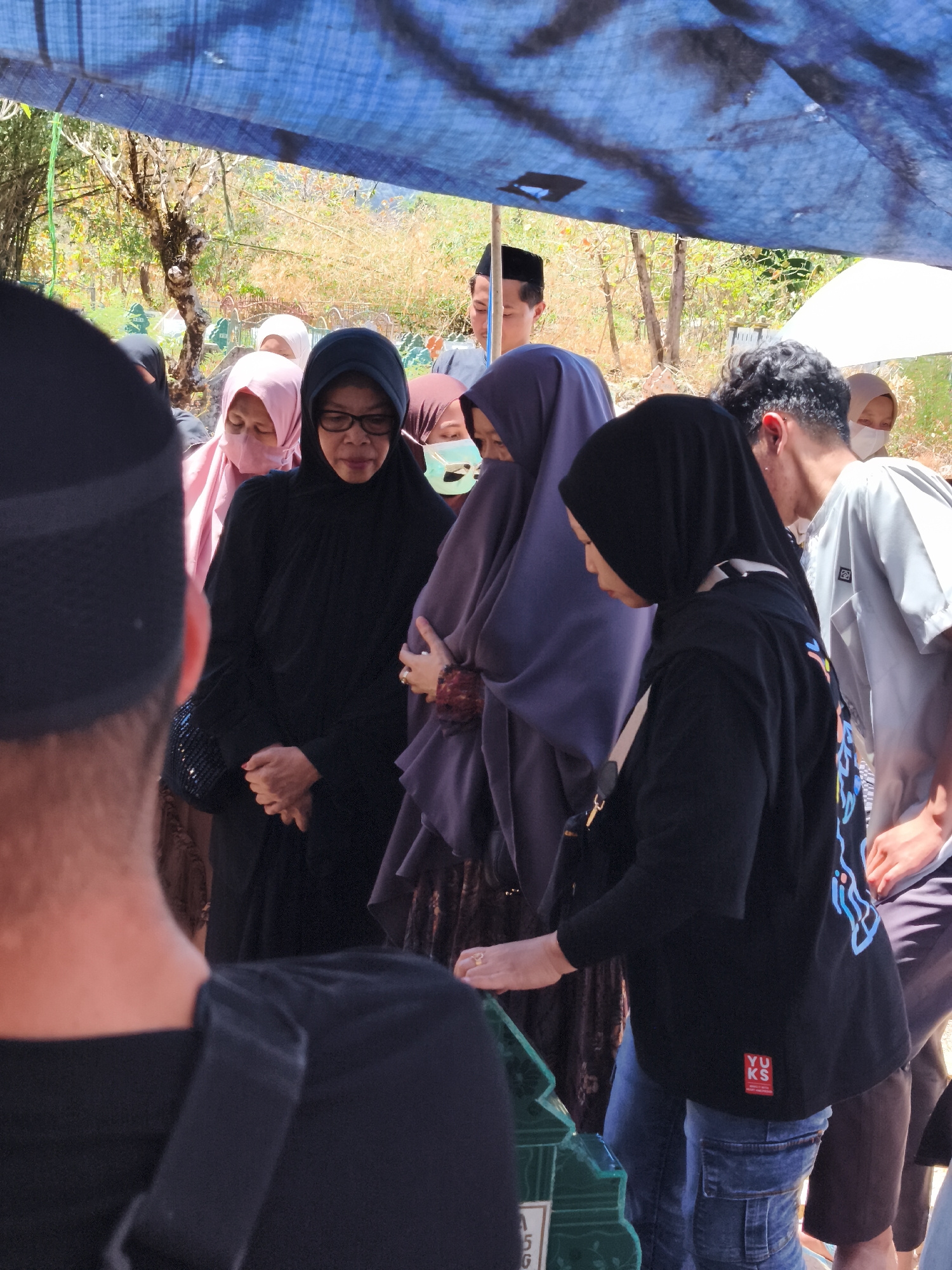 This image captures a somber gathering under a blue tarp, likely at a cemetery in Tonasa, Indonesia. A group of people, mostly women wearing hijabs, are assembled around what appears to be a grave. The central focus is on a woman in black, wearing glasses, with her hands clasped in front of her. To her right, another woman in a purple hijab holds her hand. Other individuals in the foreground and background are partially visible, some with masks on. The environment suggests an outdoor setting with trees and dry vegetation in the background. The overall mood is one of mourning and remembrance.