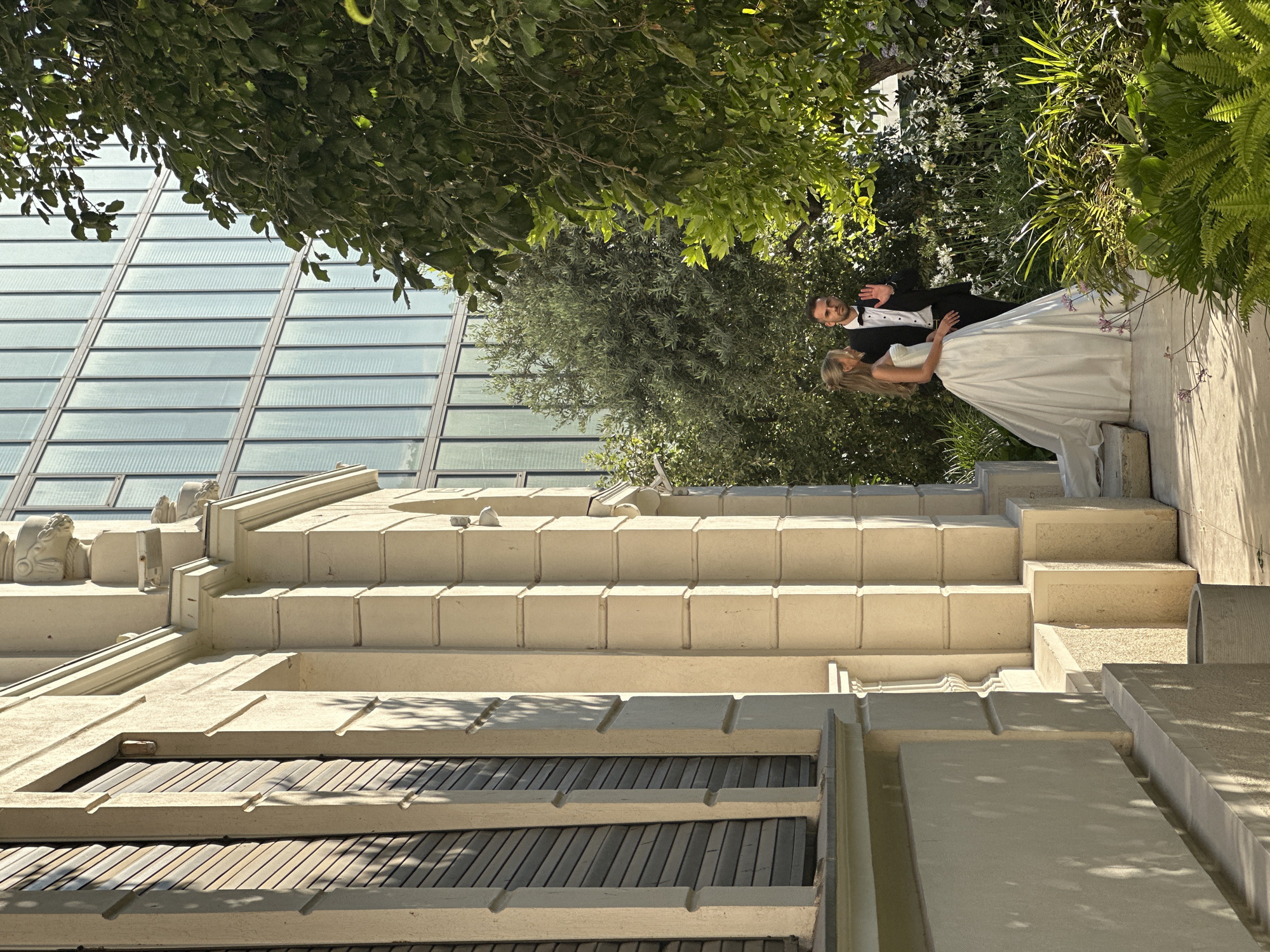 A wedding portrait of a bride and groom is captured outdoors. The groom, wearing a tuxedo with a bow tie, stands with his arm around the bride, who is dressed in a white wedding gown. They are positioned on a concrete staircase with a modern glass building in the background, partially obscured by lush green foliage. The scene appears to be set during the daytime, with sunlight casting shadows on the stairs, suggesting fair weather. The overall ambiance is romantic and celebratory, typical of a wedding event. The location appears to be Tel Aviv, Israel, indicated by the architectural style and greenery. No visible text is present in the image.