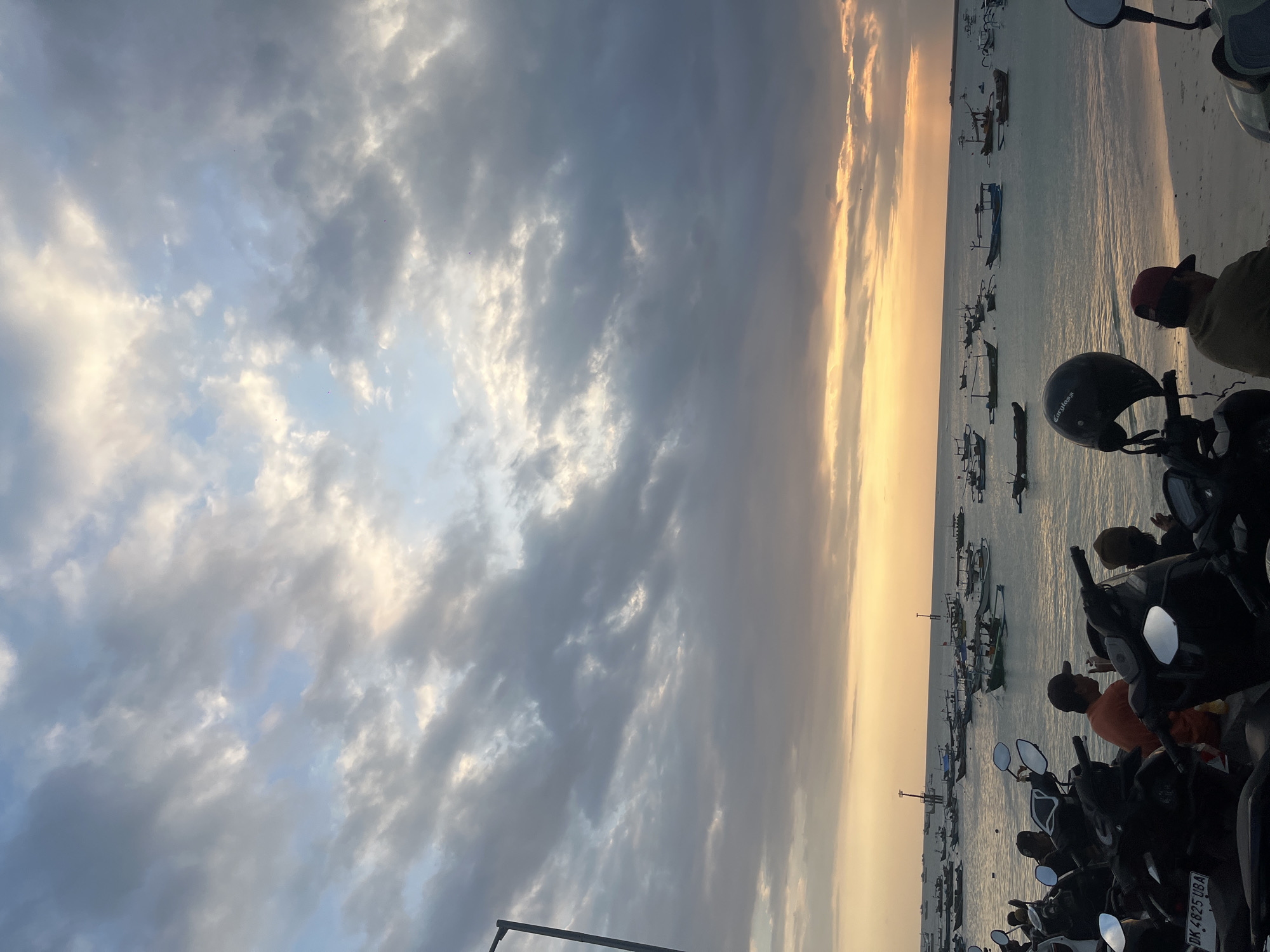 The image depicts a serene sunset scene on the coast of Kuta, Indonesia. The sky is adorned with a dramatic display of clouds, with hues of orange and yellow from the setting sun breaking through the grey. Below the sky, a tranquil body of water is dotted with numerous traditional fishing boats, their silhouettes stark against the vibrant horizon. The water meets a sandy beach in the foreground, where several motorcycles are parked. People are seen near the motorcycles, some sitting and interacting, while others are partially visible. One person is wearing an orange shirt, and another has a red baseball cap. The overall atmosphere is peaceful, capturing a moment of daily life in Kuta as the day draws to a close.
