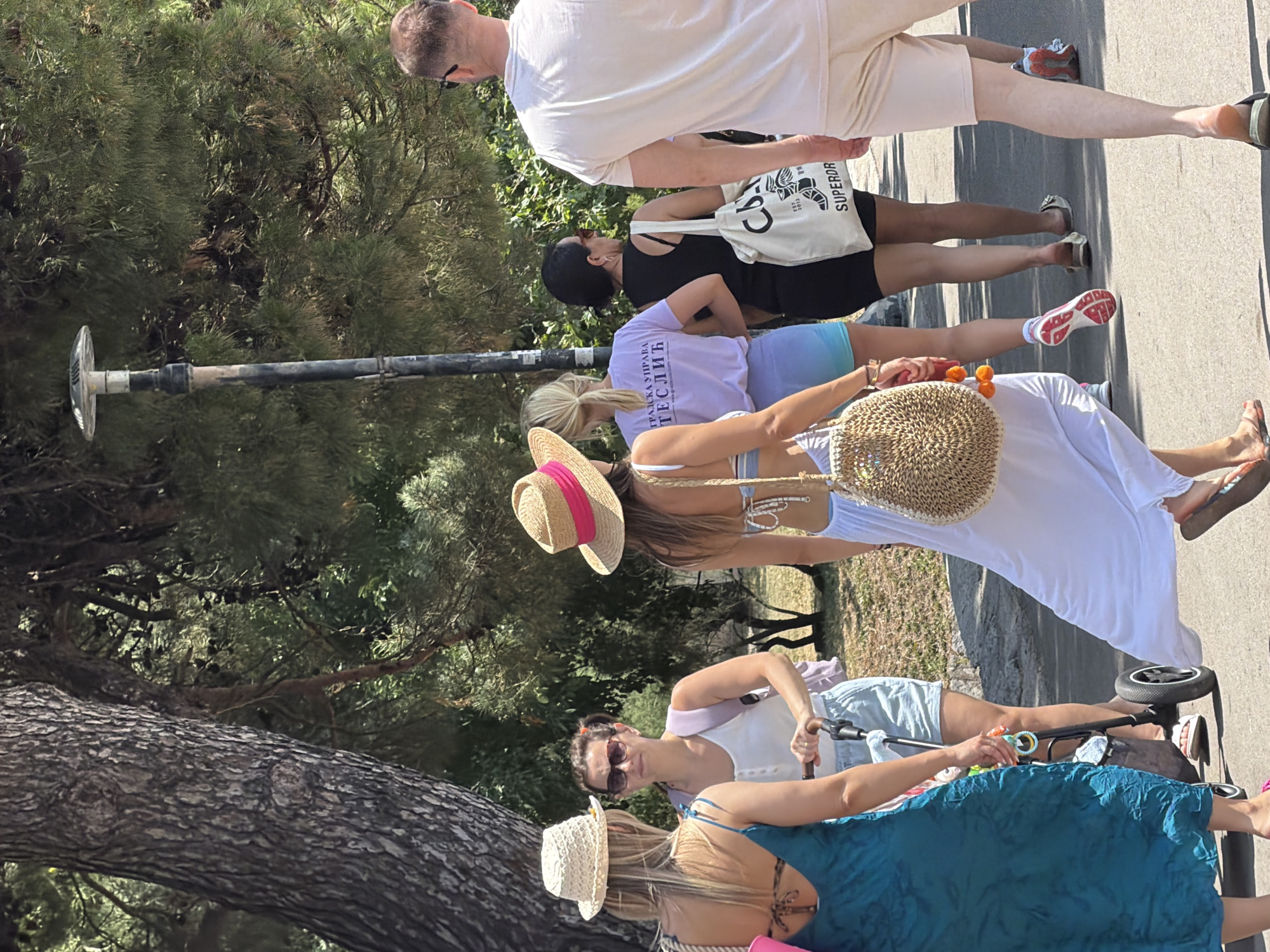 The image captures a sunny outdoor scene, likely in the city of Budva, Montenegro. The setting appears to be a pedestrian walkway or promenade, bordered by trees on the left and a paved path. Several people are walking, suggesting a leisurely stroll, possibly in a park or along the coast.
In the foreground, a woman in a turquoise dress is walking away from the camera, her back partially visible. She is wearing a straw hat and carrying a bag. Next to her, a woman with a pink backpack and sunglasses is pushing a stroller, also walking away. To her right, another woman in a straw hat with a pink ribbon and a white dress is walking with a large woven bag. Further back, a woman in a black dress is walking, carrying a white tote bag with "SUPERDRY" branding. On the far right, a man in a light-colored t-shirt and shorts is walking towards the viewer.
The environment is verdant, with lush green trees forming a canopy overhead. A lamppost is visible in the background, adding a touch of urban infrastructure to the natural setting. The overall impression is one of a pleasant day in a lively, possibly tourist-oriented, location. The presence of a stroller and casual attire suggests families and individuals enjoying outdoor activities.