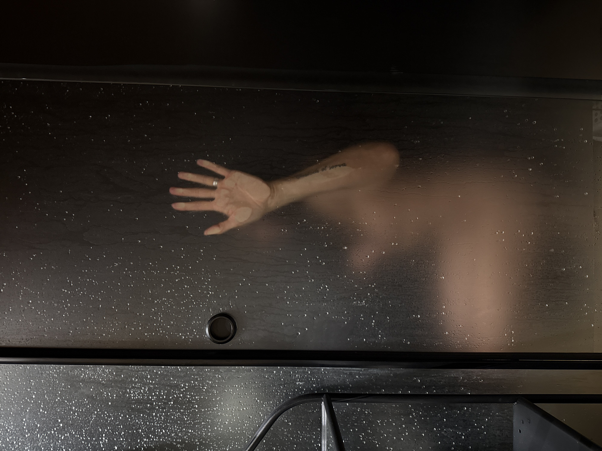 An extreme close-up shot captures a person's hand, pressed against a frosted glass shower door covered in water droplets. The hand, adorned with a ring on the index finger, is angled towards the viewer, fingers slightly spread. Beyond the glass, a blurred silhouette of a torso is visible, suggesting the presence of a person inside the shower. The left side of the image shows a section of a black spiral staircase, partially obscured by the shower enclosure. The overall scene is dimly lit, with water droplets creating a textured, abstract effect on the glass.