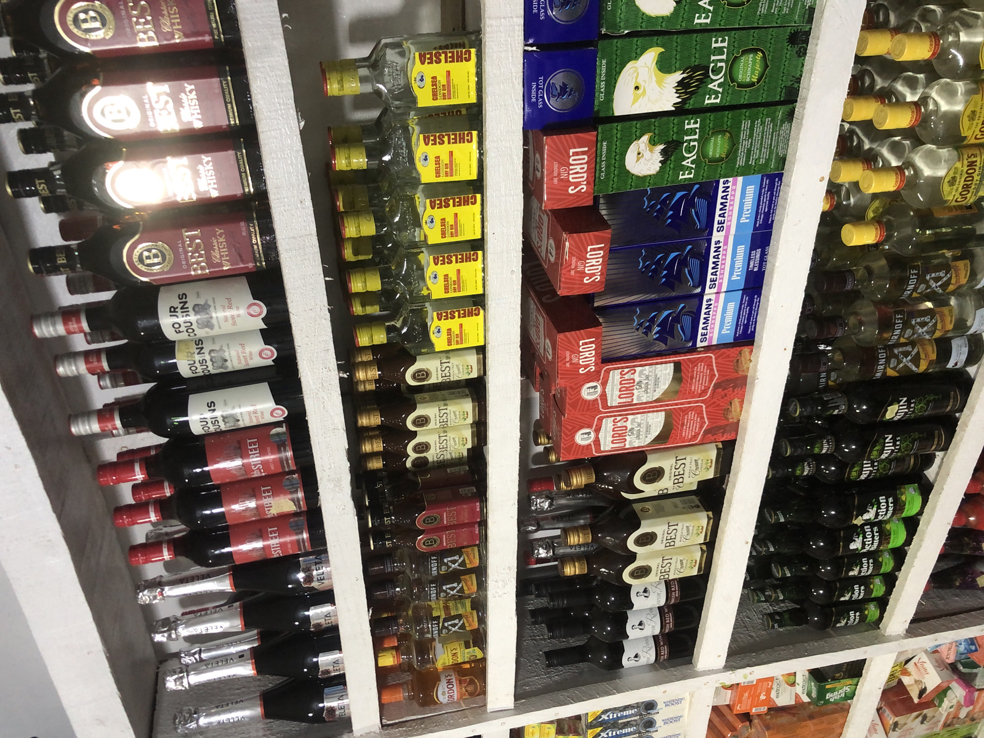 The image depicts a retail shop in Ado, Nigeria, displaying a wide assortment of alcoholic beverages on multiple shelves. The shelves are made of white wood and are stocked with various bottles and boxes of spirits, wines, and mixers.
On the top shelf, there are several bottles of wine, including red wine and what appear to be sparkling wines. Brands visible include "VELETA" and "FOUR COUSINS".
The middle shelf is heavily stocked with bottles of whisky, gin, and other spirits. Brands such as "BEST CLASSIC WHISKY", "GORDON'S", and "CHELSEA DRY GIN" are identifiable. There are also smaller bottles that appear to be miniature spirits.
The bottom shelf features more bottles of spirits, including "SMIRNOFF" vodka, and a variety of bottled bitters such as "Action Bitters" and "Bririn Bitters". Additionally, there are boxes of "LORD'S" gin and "SEAMAN'S" rum, as well as boxes featuring an eagle graphic, possibly a brand of schnapps. The shelves are well-organized, with items placed neatly in rows. The lighting appears to be artificial, likely from interior shop lights, creating some reflections on the glass bottles. The overall environment suggests a typical convenience store or liquor store setting.