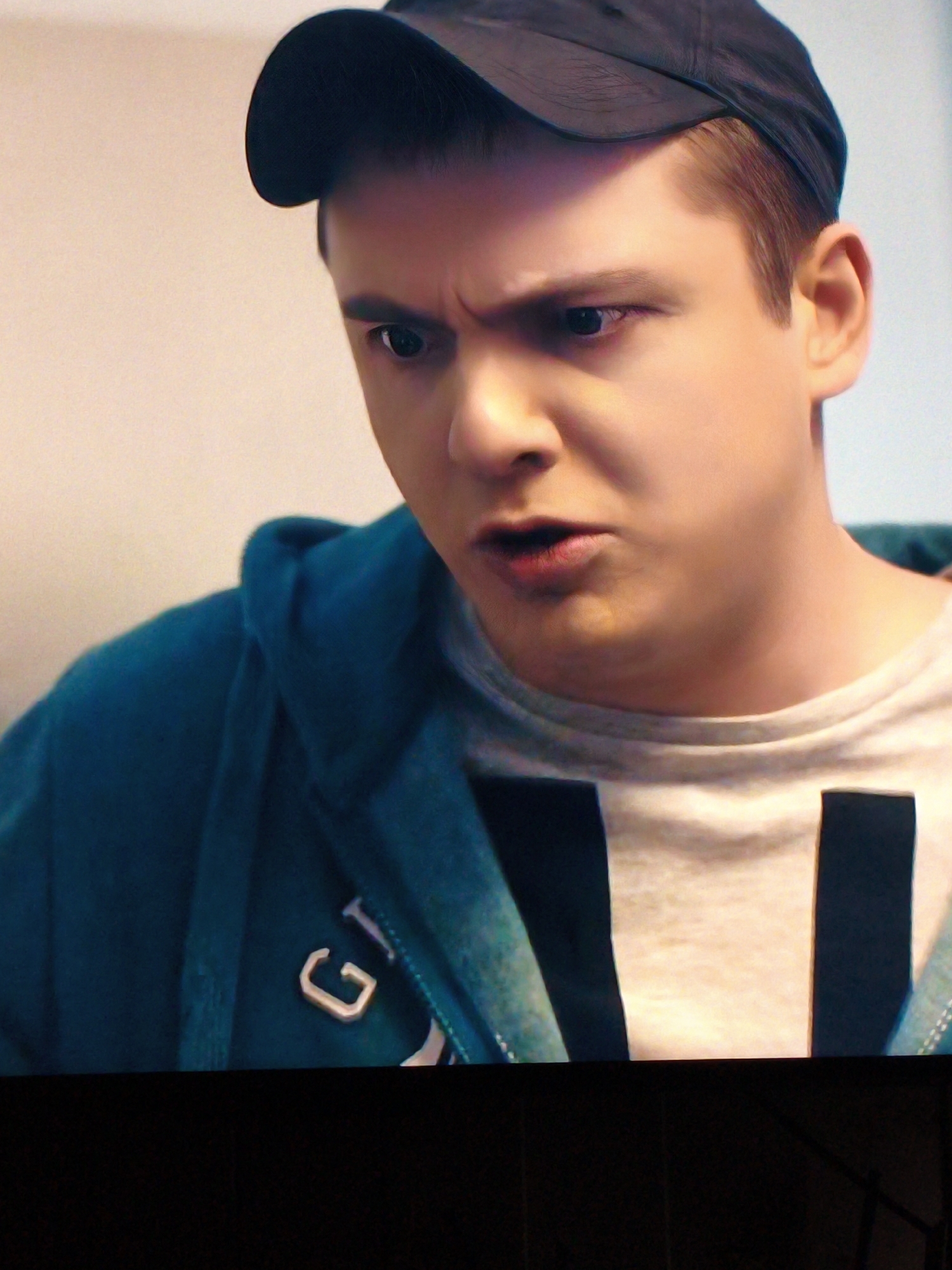 The image is a close-up, medium shot of a young Caucasian male wearing a black baseball cap and a teal hooded sweatshirt over a light-colored t-shirt with dark block lettering. The man's face is angled, with furrowed brows and his mouth slightly open as if speaking. His left eye is visible, showing a dark pupil and iris. The background is a plain, light-colored wall. The lower portion of the image is black, obscuring the man's lower body and any potential surroundings. There is no information in the image that indicates a specific location in Vladivostok, Russia.