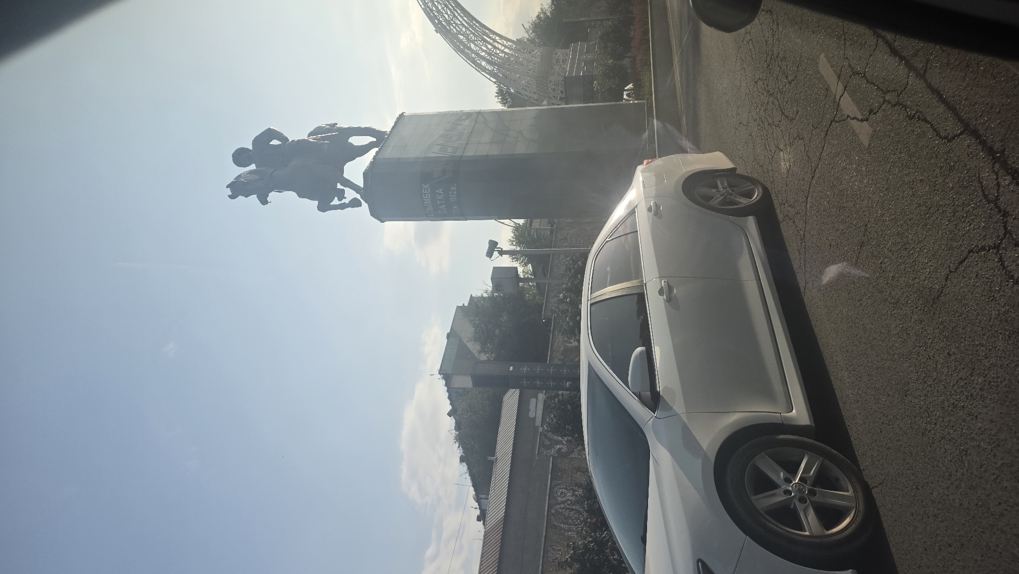 An equestrian statue of a man in a helmet stands on a tall, dark pedestal. The inscription on the pedestal reads "АЙЫМБЕК БАТКА" and "ДАТКА" followed by dates "1789-1862 г.". A white sedan is parked on the right side of the frame, facing left. In the background, there are buildings, trees, and a large arch-like structure. The sky is clear with a few clouds. The image appears to have been taken from a car moving on a paved road with visible cracks. The sun is low in the sky, casting long shadows and creating a lens flare effect. This scene is located in Osh City, Kyrgyzstan, likely near a prominent landmark or monument.