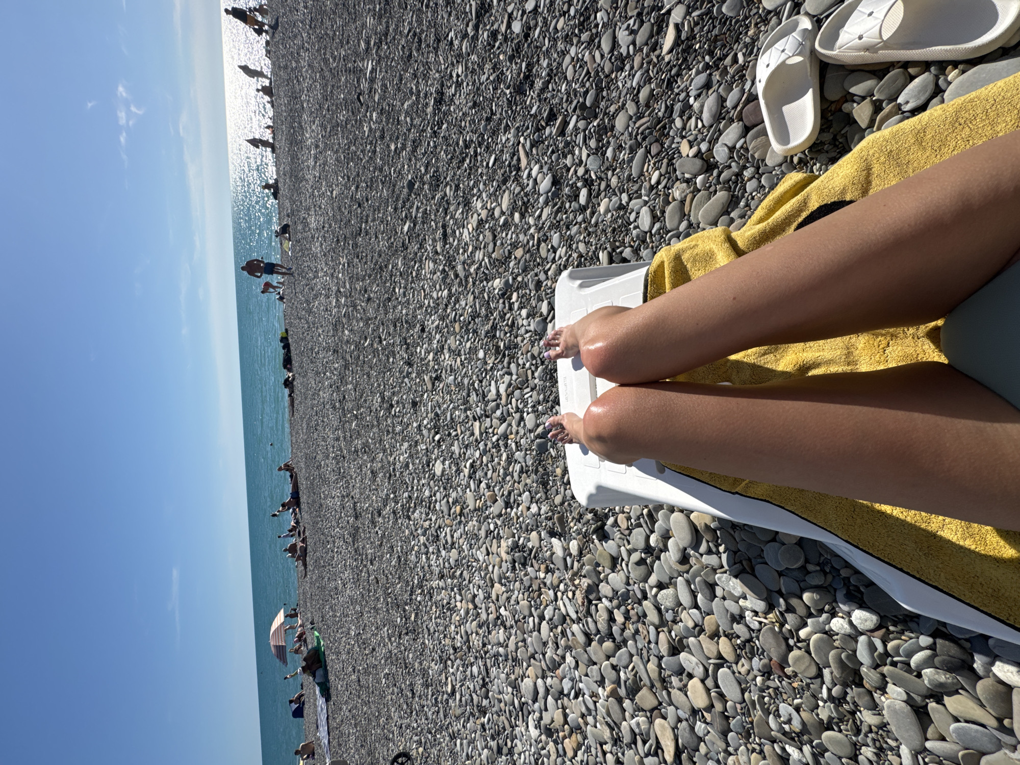 The image depicts a sunny day at a pebble beach in Sochi, Russia. In the foreground, a person's legs are stretched out on a white lounge chair, covered by a yellow towel. Their toenails are painted with purple polish. The beach is covered in grey and white pebbles, leading to the calm, turquoise sea. In the background, numerous people are scattered across the beach, some sunbathing on towels, others sitting or standing. A few individuals are wading in the shallow water. The sky is a clear blue with a few wispy clouds, and the sun's reflection shimmers on the water's surface. A beach umbrella is visible in the middle distance. White flip-flops are placed on the pebbles next to the lounge chair.