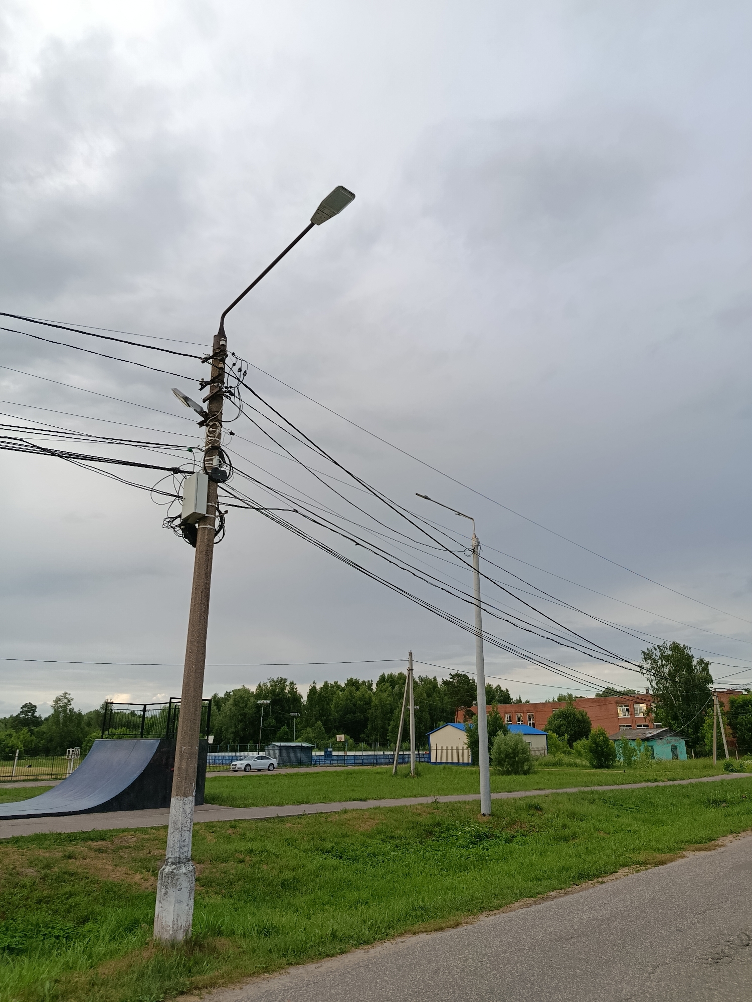 The image depicts an outdoor scene with a prominent utility pole in the foreground, laden with numerous electrical wires. To the left of the pole, a half-pipe ramp is visible, suggesting a recreational area. In the middle ground, a white car is parked near a fence, and further behind, a brick building with multiple windows can be seen. The background consists of a dense line of trees under a cloudy sky. The overall environment suggests a semi-urban or suburban setting.