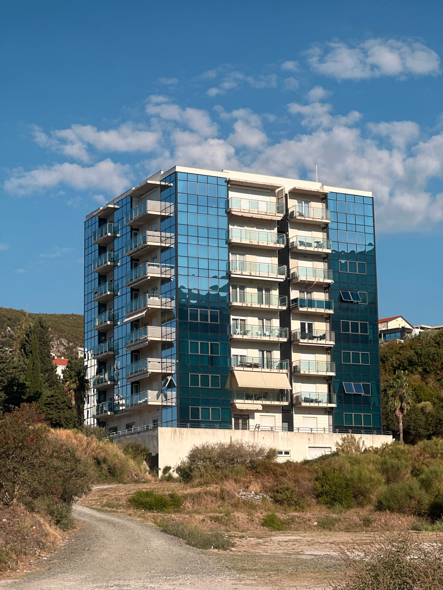 A tall, modern building with a blue glass facade and balconies stands on a hillside in Boreti, Montenegro. The building has at least ten stories, with each floor featuring several balconies with glass railings. The blue glass reflects the sky and clouds, creating a striking visual effect.
The building is surrounded by lush greenery, including trees and shrubs, typical of the Mediterranean landscape. A dirt path winds its way up the hillside towards the building. The sky above is a vibrant blue, dotted with fluffy white clouds. The overall impression is one of modern architecture harmoniously integrated with the natural environment.