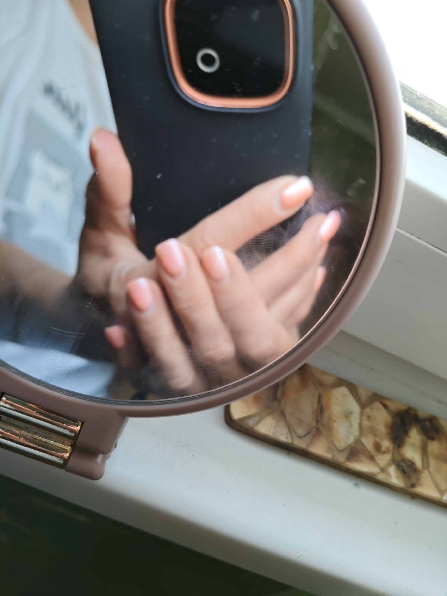 A close-up, slightly angled shot shows a hand reflected in a handheld mirror. The hand, which is the primary subject, has neatly manicured fingernails painted a pale peach color. The reflection is of the person holding the mirror, with their hand positioned in front of a dark-colored phone case, which has a circular camera lens with a copper-colored rim. The mirror itself is round and has a light pink frame. The background visible in the reflection shows a blurred white surface and a hint of green. To the right of the mirror, on a white windowsill, is a decorative object made of tessellated natural materials, resembling shell fragments, in shades of beige and brown. The overall scene is indoors, suggesting a personal setting.