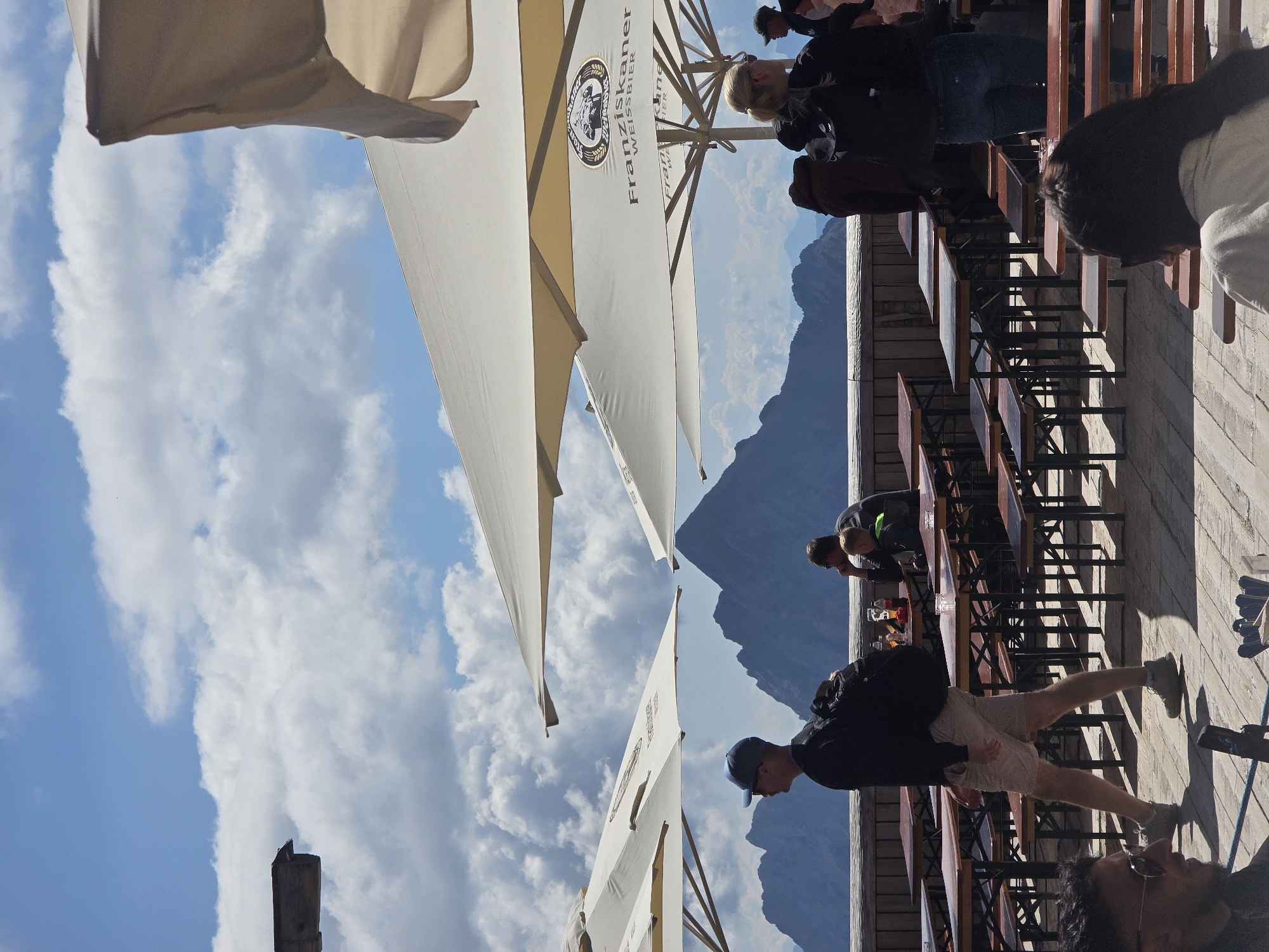 The image depicts an outdoor seating area with several people enjoying the view of the Bavarian Alps, likely at the summit of the Zugspitze mountain, near Garmisch-Partenkirchen, Germany. Several large, cream-colored umbrellas provide shade over rows of wooden tables and benches. A man in a black shirt, beige shorts, and a blue baseball cap walks across the wooden deck, carrying a backpack. In the background, other individuals are seated at tables. The majestic mountain range forms a striking backdrop under a bright blue sky dotted with fluffy white clouds. The scene suggests a leisurely atmosphere with tourists taking in the scenic beauty.