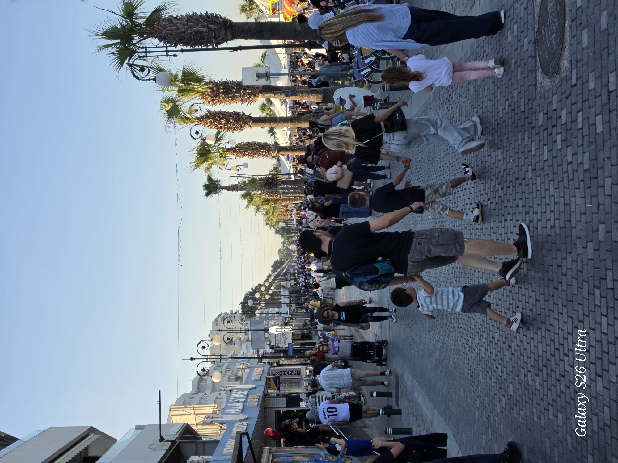 The image shows a pedestrian street in Larnaca, Cyprus, packed with people. The street is lined with palm trees and ornate street lamps, and buildings with shops on the ground floor. Many people are walking along the paved sidewalk, including families with children. A man wearing a white jersey with the number 10 is visible in the foreground. The sky is clear and blue, indicating a sunny day.