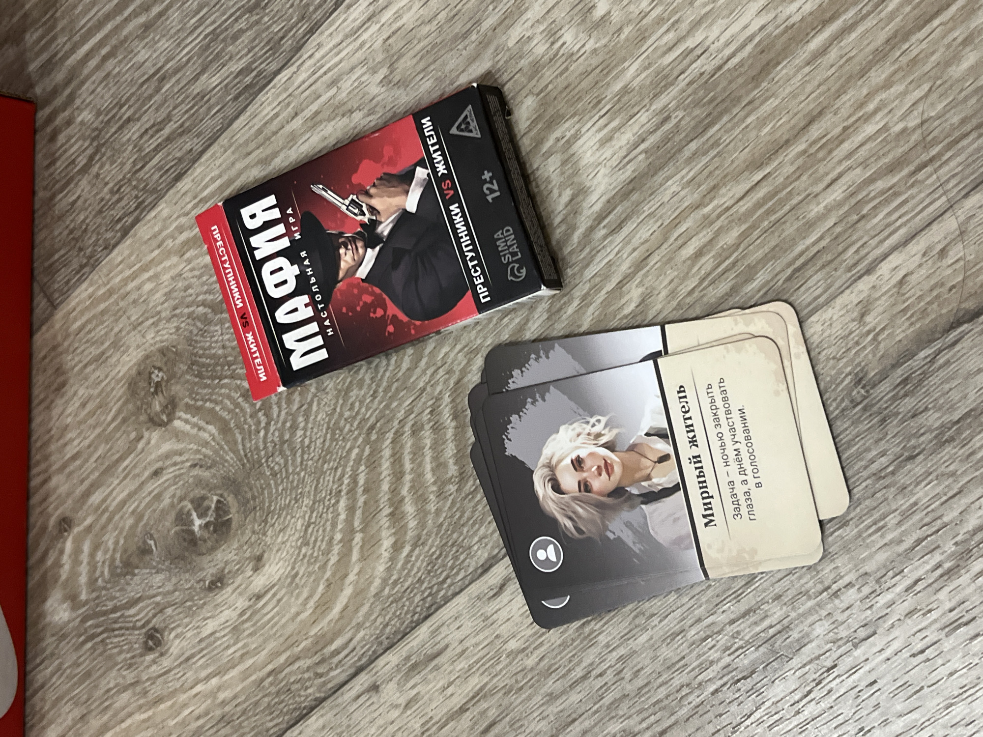 This is a high-angle, eye-level shot of a board game and cards on a wooden floor. The board game box, titled "МАФИЯ" (Mafia) in large white letters, features a graphic of a man in a fedora and suit holding a pistol. Text on the box reads "НАСТОЛЬНАЯ ИГРА" (Board Game) and "ПРЕСТУПНИКИ VS ЖИТЕЛИ" (Criminals vs. Citizens). Below this, "SIMA LAND" is printed alongside a 12+ age rating. A red border frames the top and right edges of the box.
Scattered to the left of the box are several cards from the game. The top visible card displays an illustration of a young woman with blonde hair and a white shirt. Text on this card reads "Мирный житель" (Peaceful Resident) and describes the character's role: "Задача – ночью закрыть глаза, а днём участвовать в голосовании." (Task - close eyes at night, and participate in voting during the day). Other cards are stacked beneath, partially obscured. The wooden floor appears to be a laminate with a light brown, grayish wood grain pattern. The scene does not contain any elements indicating a specific location within Дачное, Russia.