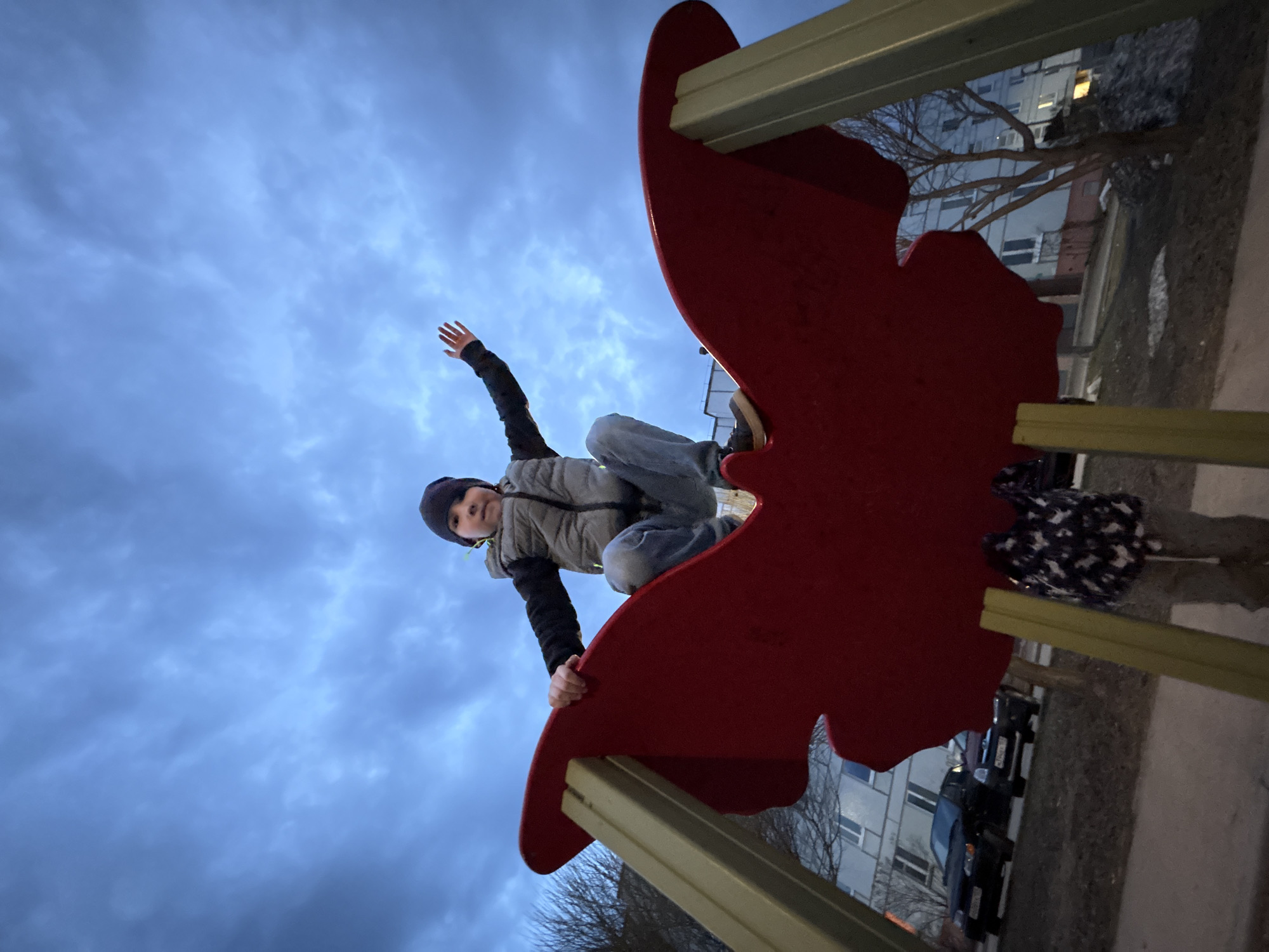 A low-angle, slightly rotated shot depicts a child perched atop a red, butterfly-shaped playground structure in an outdoor setting. The child, wearing a dark beanie, a gray vest over a black long-sleeved shirt, and jeans, is positioned centrally in the frame. The child has one arm raised, with the other gripping the edge of the structure. Another child, wearing patterned clothing, stands off to the side and partially out of view. The sky, with prominent cloud cover, dominates the background. Buildings, including a multi-story residential building, are partially visible behind trees with bare branches. Dark colored cars are seen near the residential building. Based on visual cues and known landmarks, the location is consistent with the city of Chaikovsky, Russia, likely in a playground or recreational area. The photo appears to have been taken during dusk or dawn, given the lighting conditions.