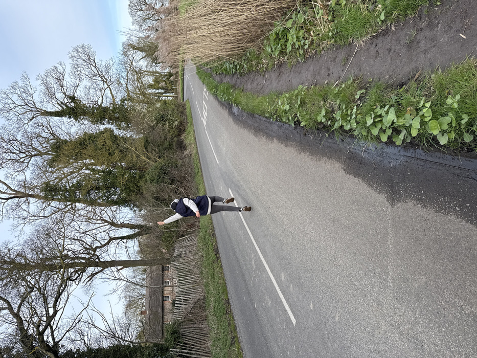 An individual is captured mid-stride, running along a rural asphalt road in North Norfolk, United Kingdom, under an overcast sky. The person, viewed from behind, wears a dark blue sleeveless vest over a light-colored long-sleeved top, grey athletic leggings, and brown running shoes. White over-ear headphones are visible. Their right arm is raised vertically, hand partially clenched, while the left arm is positioned lower and bent.
The road features a white dashed center line and the word "SLOW" painted in white ahead of the runner. To the left of the road, a light-colored wooden picket fence borders dense vegetation, consisting primarily of bare deciduous trees and some evergreens. A traditional thatched cottage with white window frames is visible through the trees in the background. To the right of the road, a narrow dirt verge with green ground cover transitions into a thick stand of tall, dry, light brown reeds. The scene suggests a late autumn or winter environment.