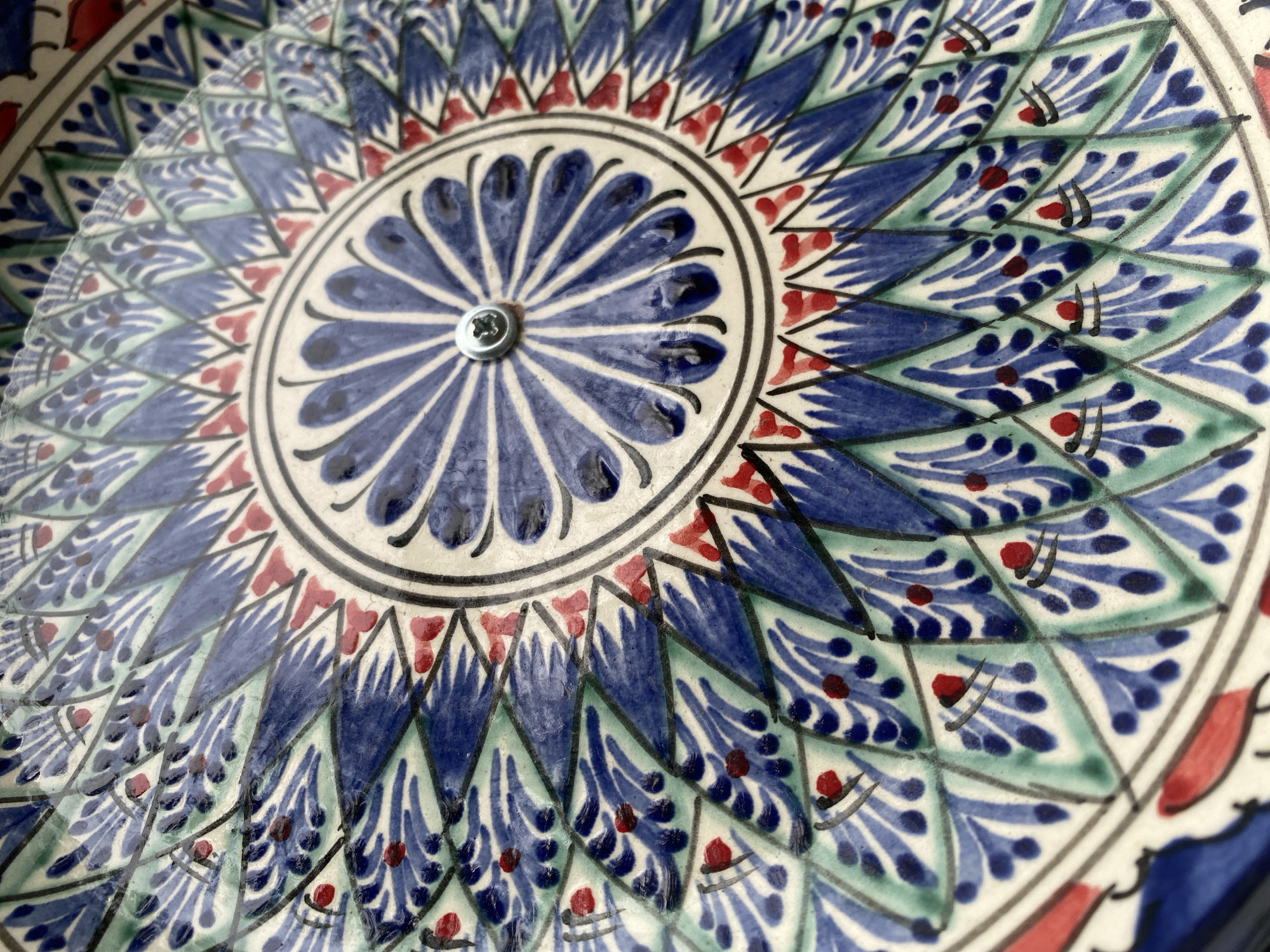 This media file is a close-up, overhead shot of a decorative ceramic plate, likely part of a set. The plate features a complex, radial pattern in shades of blue, green, and red, with black accents. The central motif is a starburst of blue, petal-like shapes radiating outwards, all centered around a small, silver screw.
Surrounding the central motif, the design becomes more intricate. Alternating sections of blue and green diamonds are filled with delicate, feather-like blue patterns, punctuated by small red dots and subtle black markings that resemble small strokes or leaves. Red triangular elements form an outer ring of the design, with larger blue petal-like shapes continuing the radial theme towards the edge of the plate. The background of the plate is off-white, with a slight texture.
No people, activities, or specific events are depicted. The focus is solely on the intricate artistry of the ceramic piece. There is no visible text. The image's lighting suggests it was taken indoors under natural or artificial light, and there are no environmental cues that indicate specific weather or time of day. The context provided places this object in Almaty, Kazakhstan, suggesting it may be a piece of local craftsmanship. The overall impression is one of detailed and colorful decorative artistry.