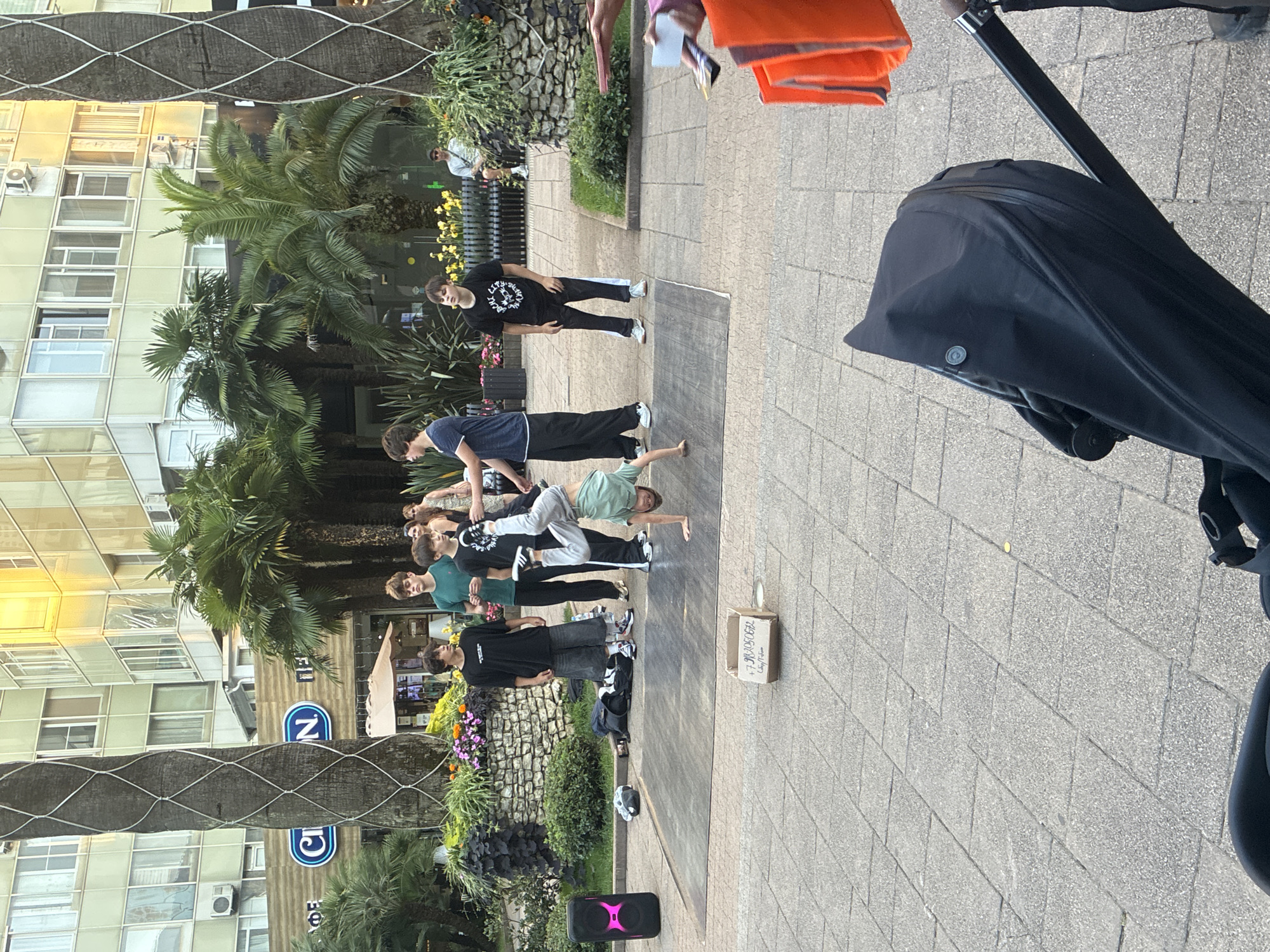 This image depicts an outdoor scene on a paved plaza, likely in Sochi, Russia, characterized by palm trees and a modern building with numerous windows. The focus is on a group of young males engaged in breakdancing. One individual is performing a handstand on a portable dance mat, while others stand around, observing or waiting their turn. To the left of the dance mat, a portable speaker with pink lighting is positioned. A cardboard box sits on the ground near the dance mat, possibly for donations, with some writing on it. In the background, more palm trees and lush greenery are visible, along with benches and a building. In the foreground, a black stroller is partially visible, suggesting the presence of a family or onlookers. The overall atmosphere appears to be a public space where street performers are entertaining.