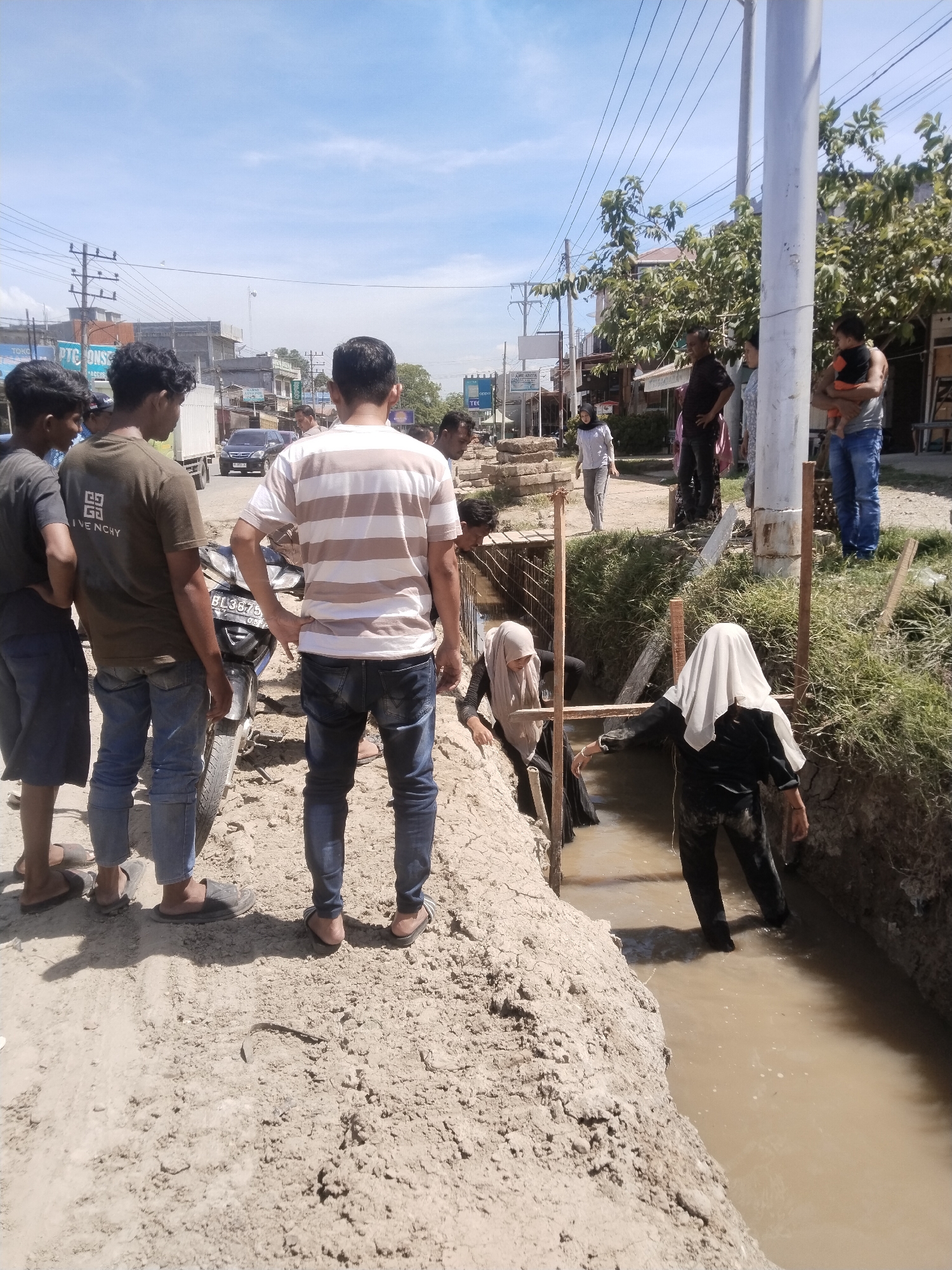 In Manetunong, Indonesia, a group of people are gathered at a construction site. Some stand on the edge of a newly dug trench filled with muddy water. Two women, wearing headscarves, are inside the trench, seemingly working on something. Other people are watching from a short distance away, including a person holding a baby. A motorcycle is parked near the edge of the trench where the standing group is located. In the background, there's a street with buildings and power lines under a partly cloudy sky.