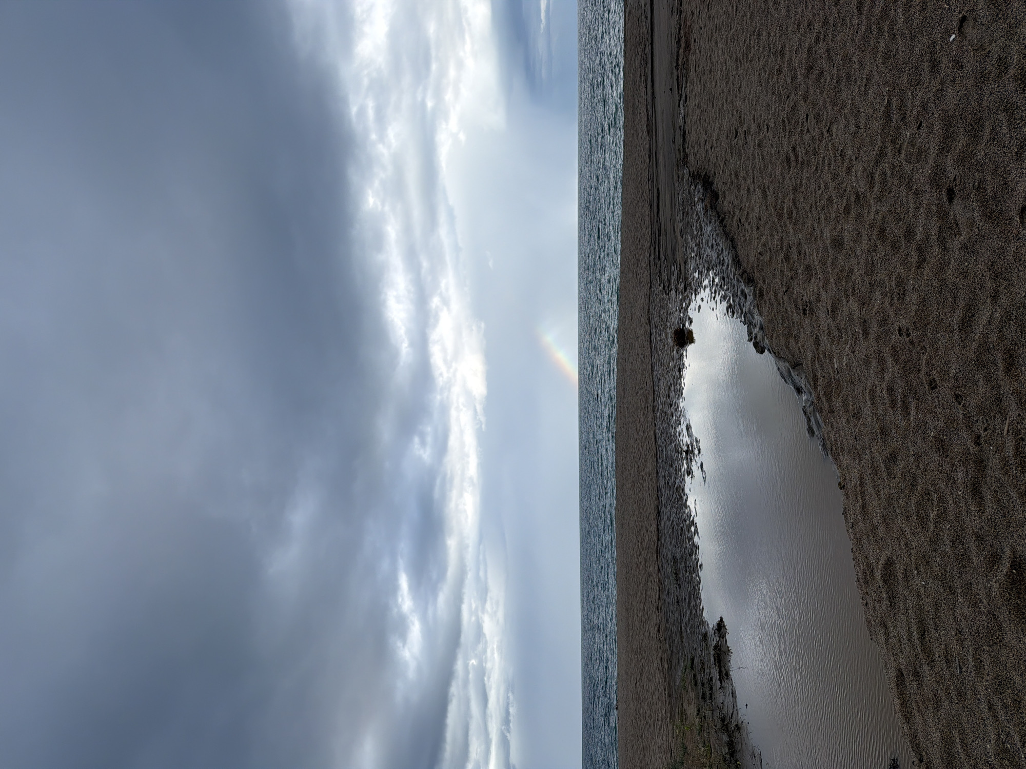 A view of Sa Punta, Spain featuring the beach and sky. A large pool of water occupies the lower right, reflecting the sky. The beach extends along the right, composed of dark, grainy sand. Next to the beach is the sea, and the sky and clouds above. A fragment of a rainbow appears in the sky.