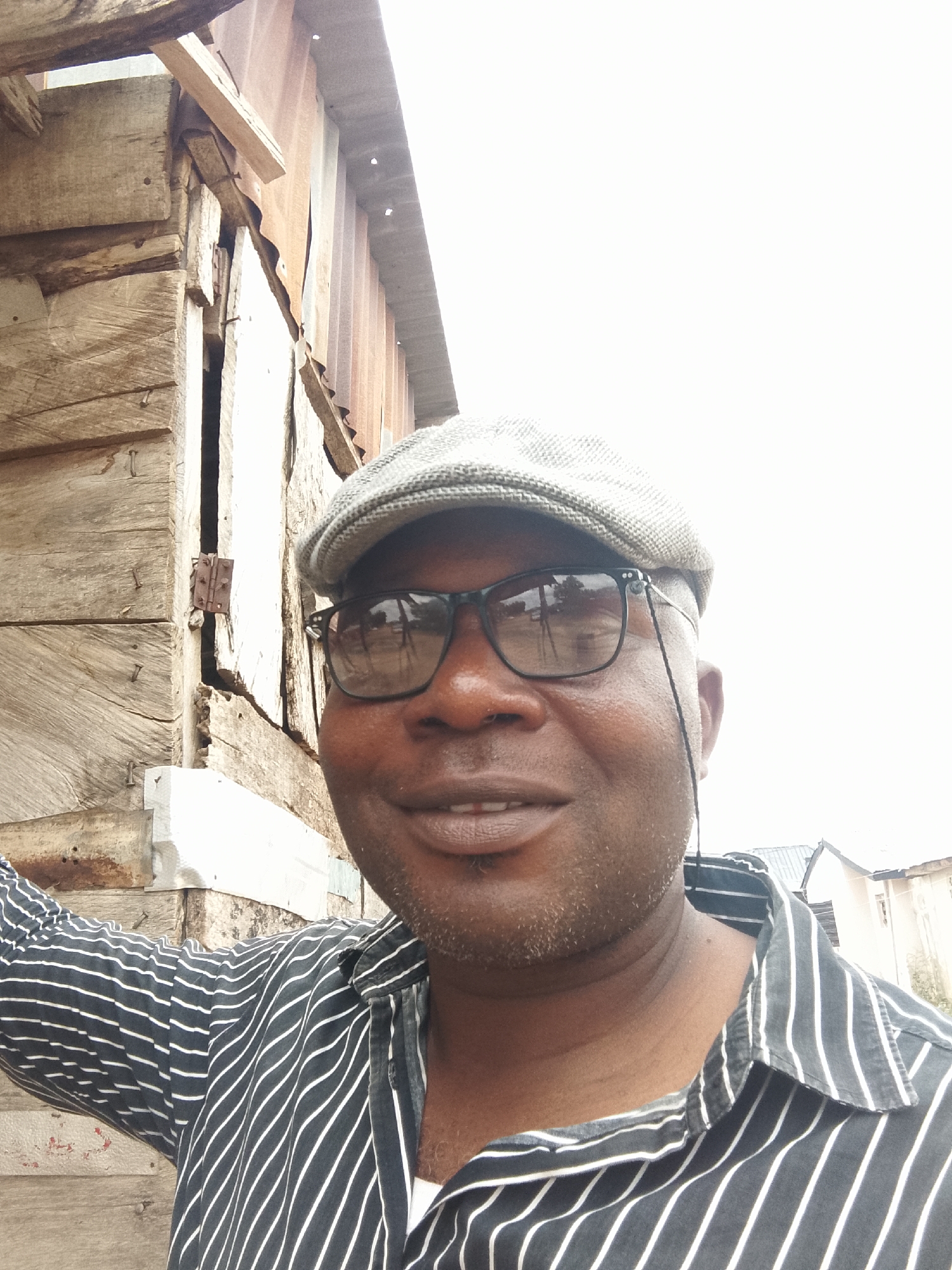 A medium shot depicts a smiling, dark-skinned man wearing a newsboy cap and glasses, standing next to a wooden structure with corrugated metal roofing in an urban setting. The man is wearing a black and white striped shirt. The wooden structure appears weathered and roughly constructed. A building is visible in the background. The location is unknown.