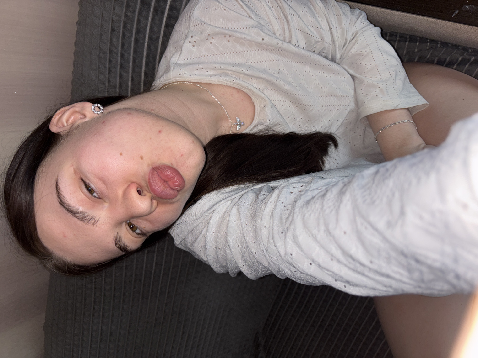 A young woman with dark hair is lying on a grey, striped couch. She is wearing a white, eyelet-style top and a silver cross necklace and a silver bracelet. She has an earring in her left ear. Her face is angled towards the camera, and she is puckering her lips. The location is not discernible from the image.