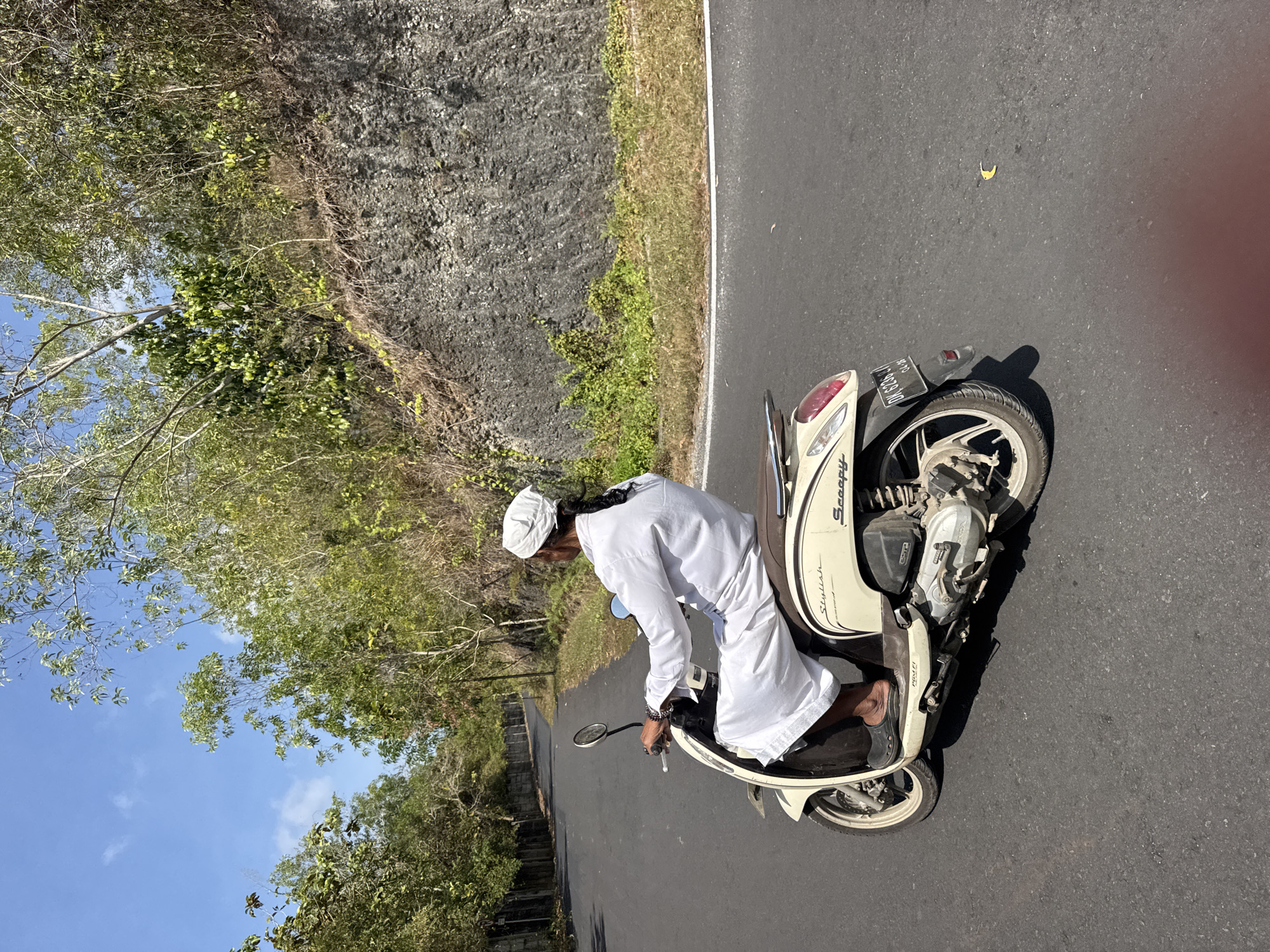 A person wearing a white head covering and white traditional attire rides a cream-colored Honda Scoopy scooter on a paved road. The person is viewed from behind and to the right, with their face turned away from the camera. The scooter has a black license plate with white text that reads "DK 6236 SJ". The road is bordered by a dry, rocky embankment with sparse vegetation on the right and trees with a concrete wall on the left. The sky is blue with scattered clouds. The scene appears to be outdoors in a rural or semi-rural area, potentially in Kuta Selatan, Indonesia.