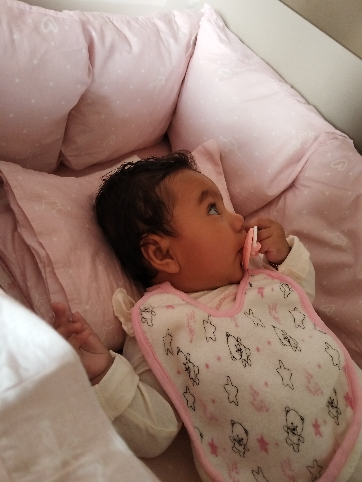 A baby lies on pink bedding with heart and star patterns. The baby has dark hair, is wearing a bib with bear and star patterns and a pink trim, and has a pink pacifier in its mouth. The location is Unknown, Unknown.